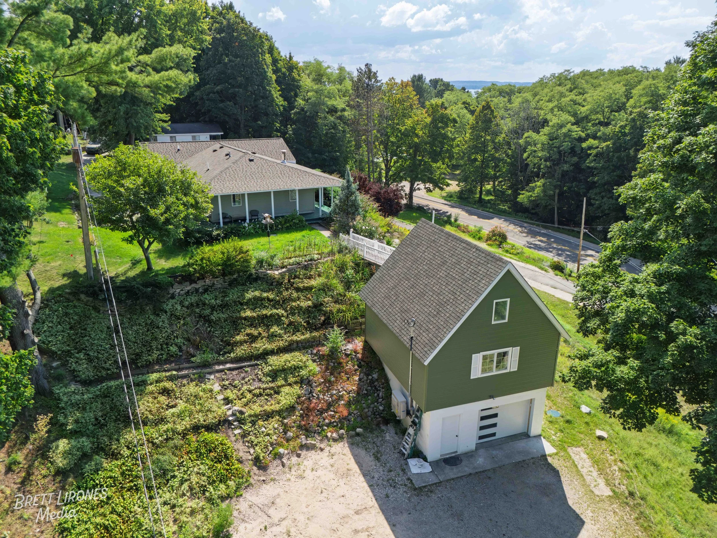A green house with white trim and a garage at the bottom, situated on a gravel driveway. A ladder leans against the garage. There are multiple trees and landscaped gardens surrounding the house. In the background, there is a road and a wooded area, w