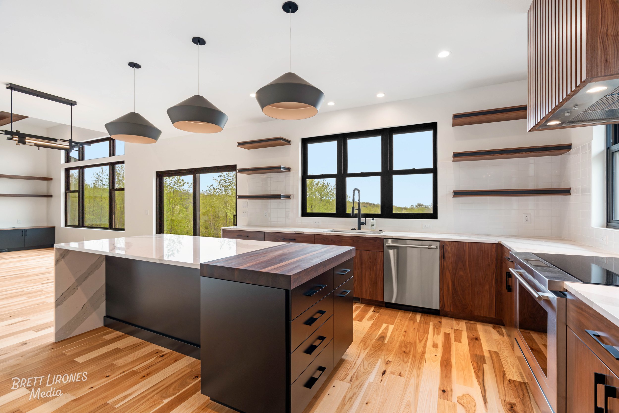 Modern kitchen with wood flooring, black-framed windows, white countertops, and a kitchen island with black base and wooden top, pendant lights, and open shelving.