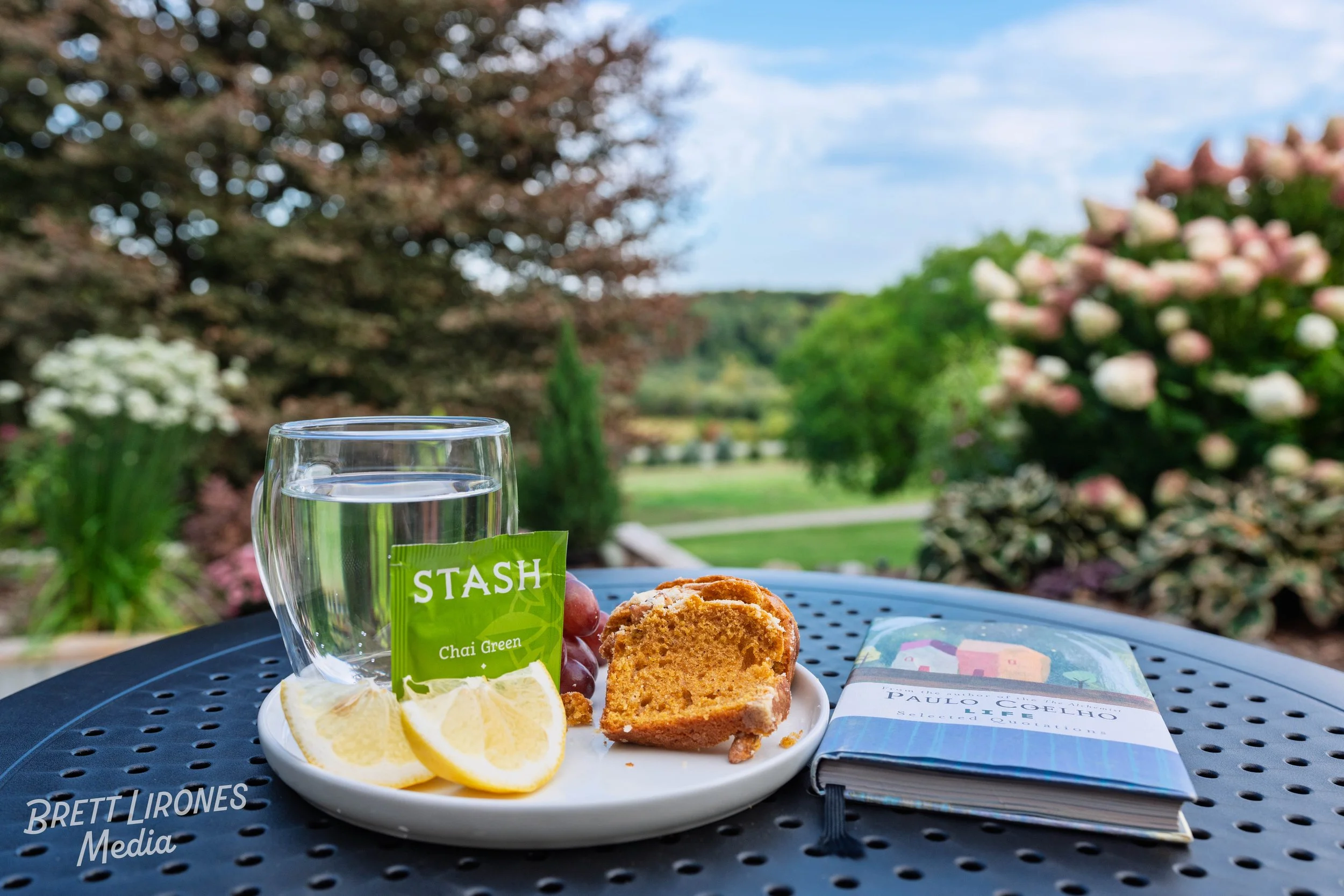 A glass of water, a lemon wedge, grapes, a slice of banana nut bread, a box of Stash chai green tea, and a book titled 'Paulo Coelho' on a black outdoor table with a garden and trees in the background.