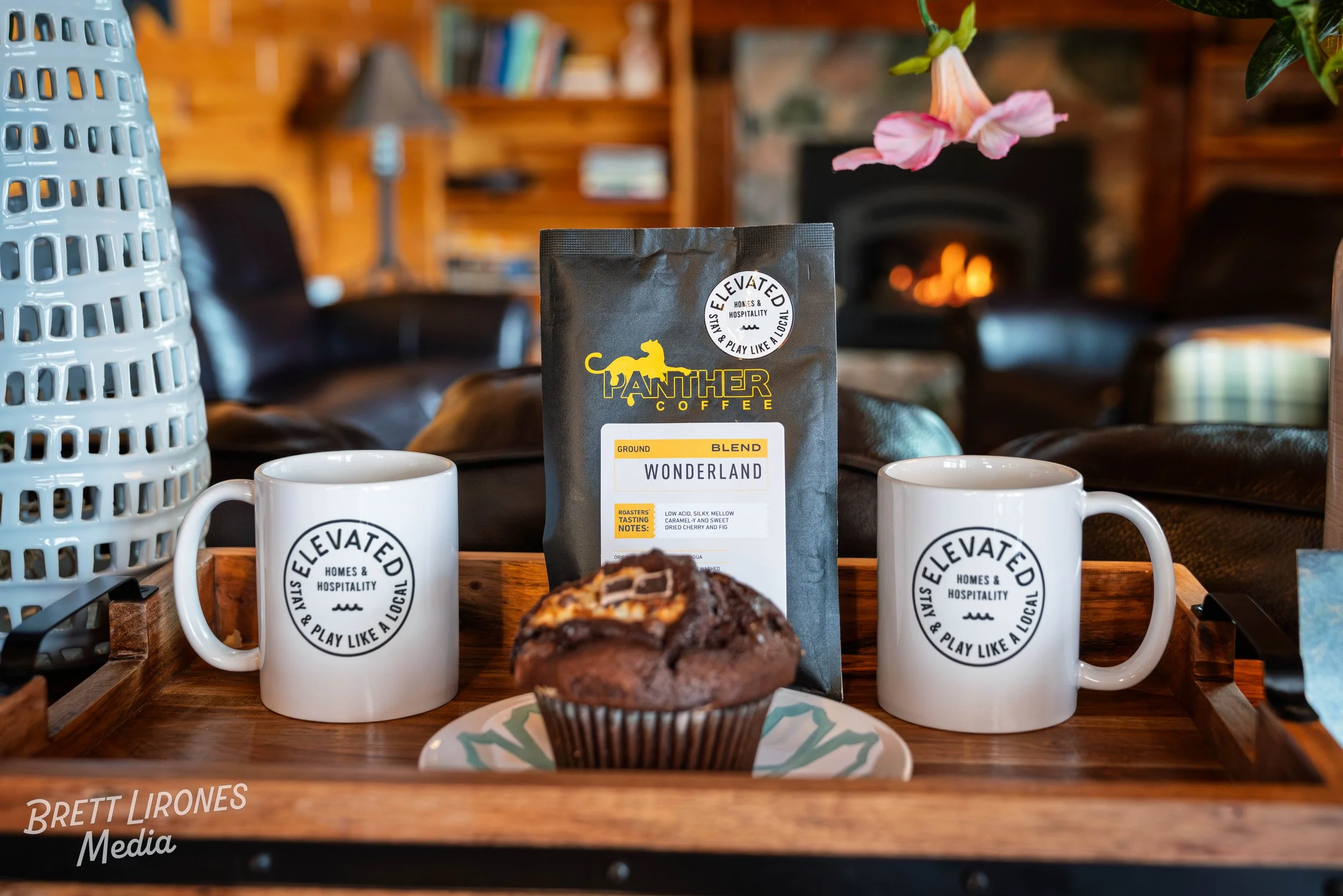 A tray with two white mugs that have a black circle logo reading 'Elevated Homes & Hospitality, Stay & Play Like a Local', a muffin with chocolate and caramel on a decorative plate, and a bag of Panther Coffee Wonderland blend coffee in a cozy room w