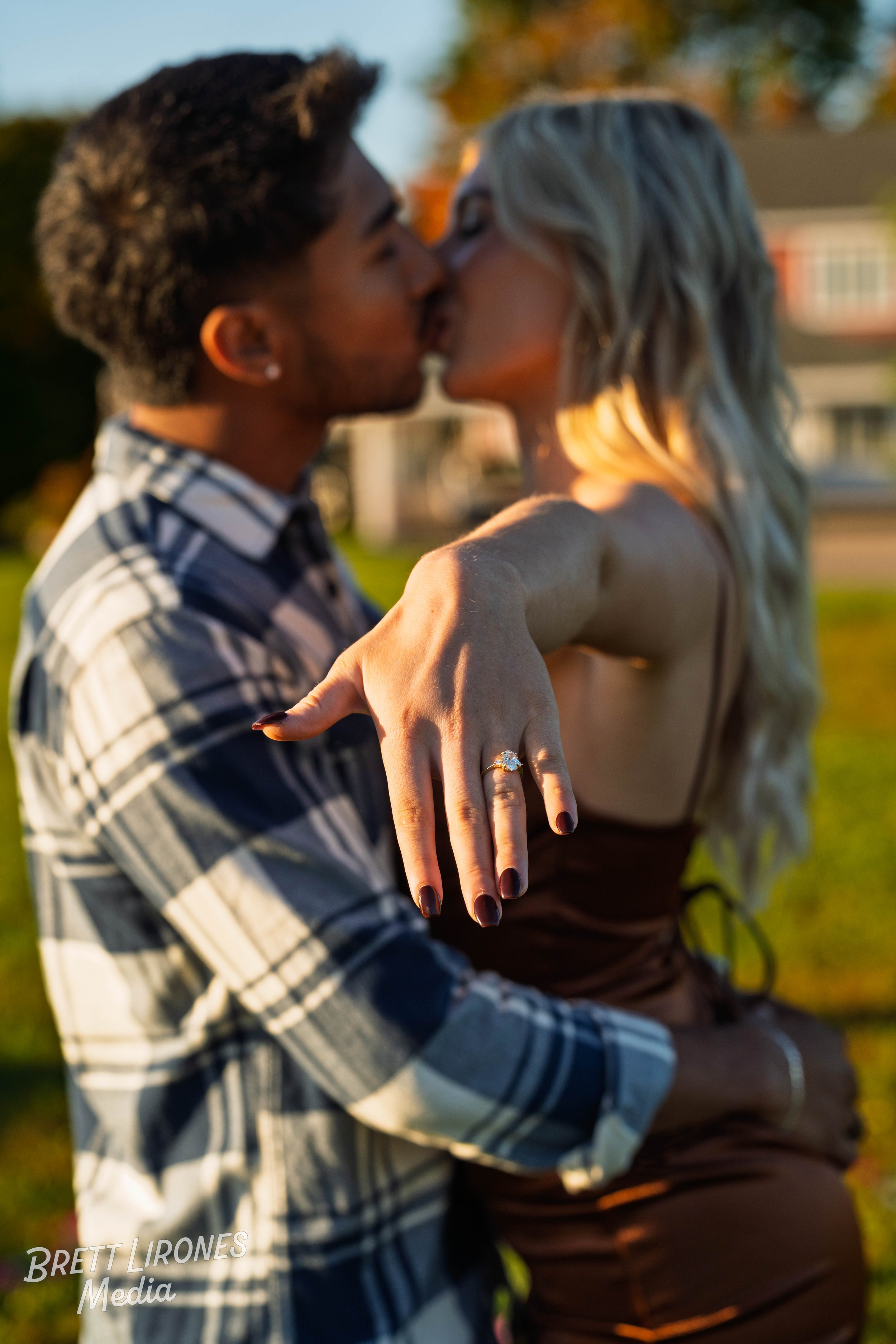 A couple kissing outdoors during golden hour, showing a close-up of the woman's hand with a diamond engagement ring.