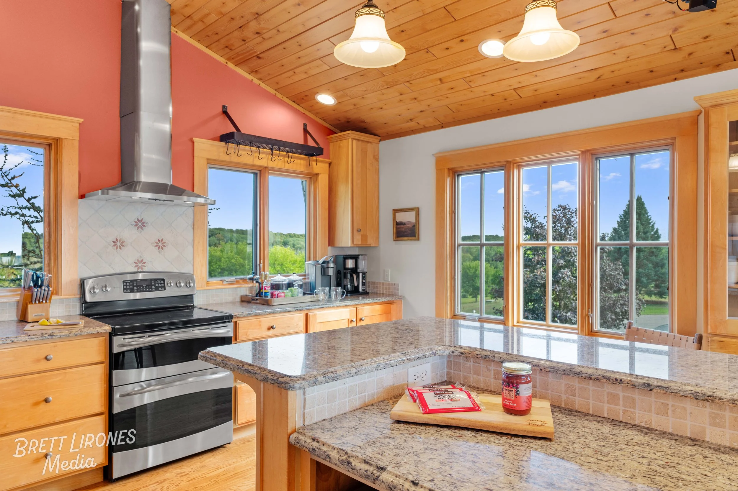 Kitchen with granite countertops, wooden cabinets, stainless steel stove and range hood, coffee maker, large windows showing green trees outside, wooden ceiling with pendant lights, and a small cutting board with a jar of jam on the counter.