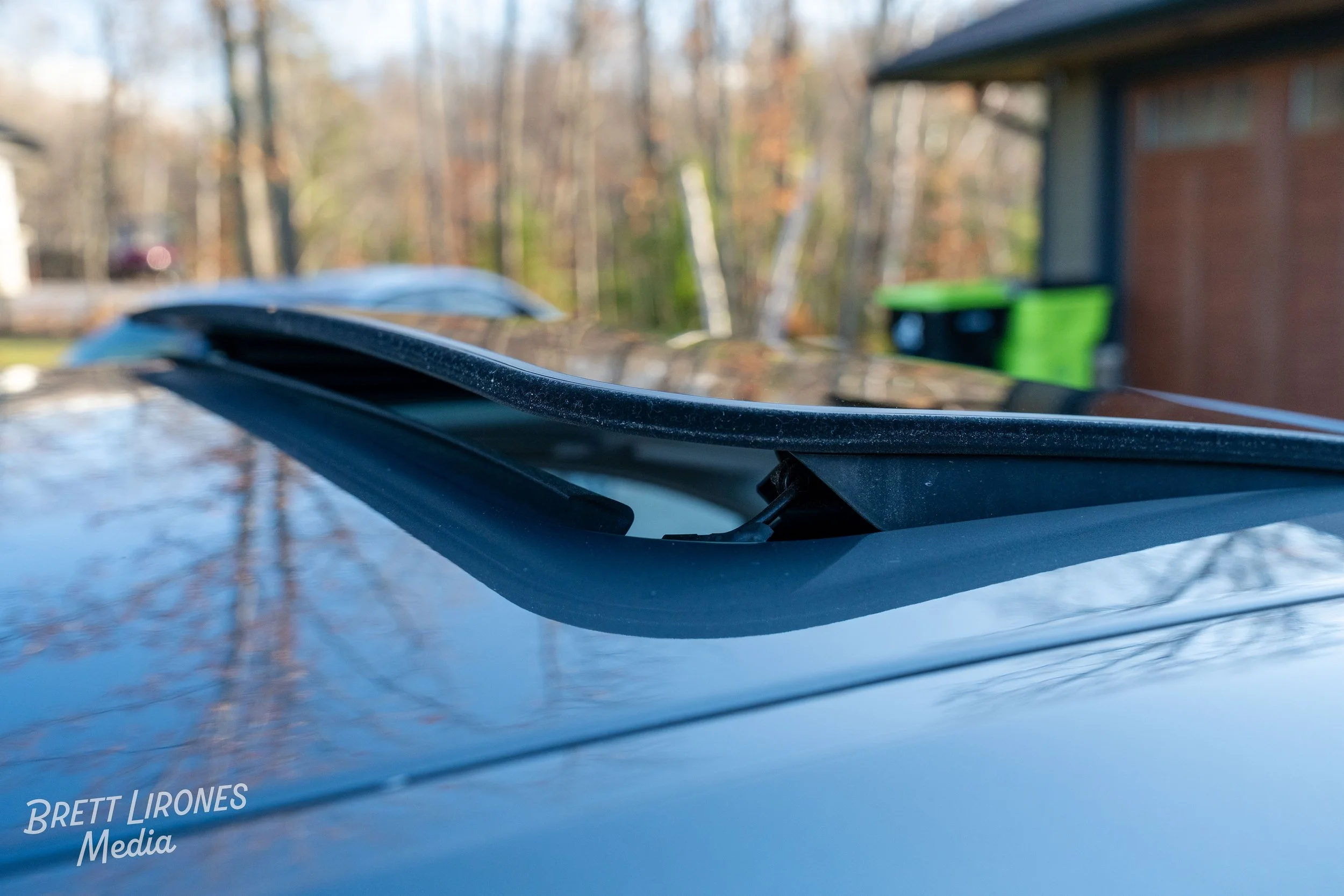 Close-up of a car windshield wiper on a shiny car hood with a blurred background of trees, a brick building, and a green trash bin.