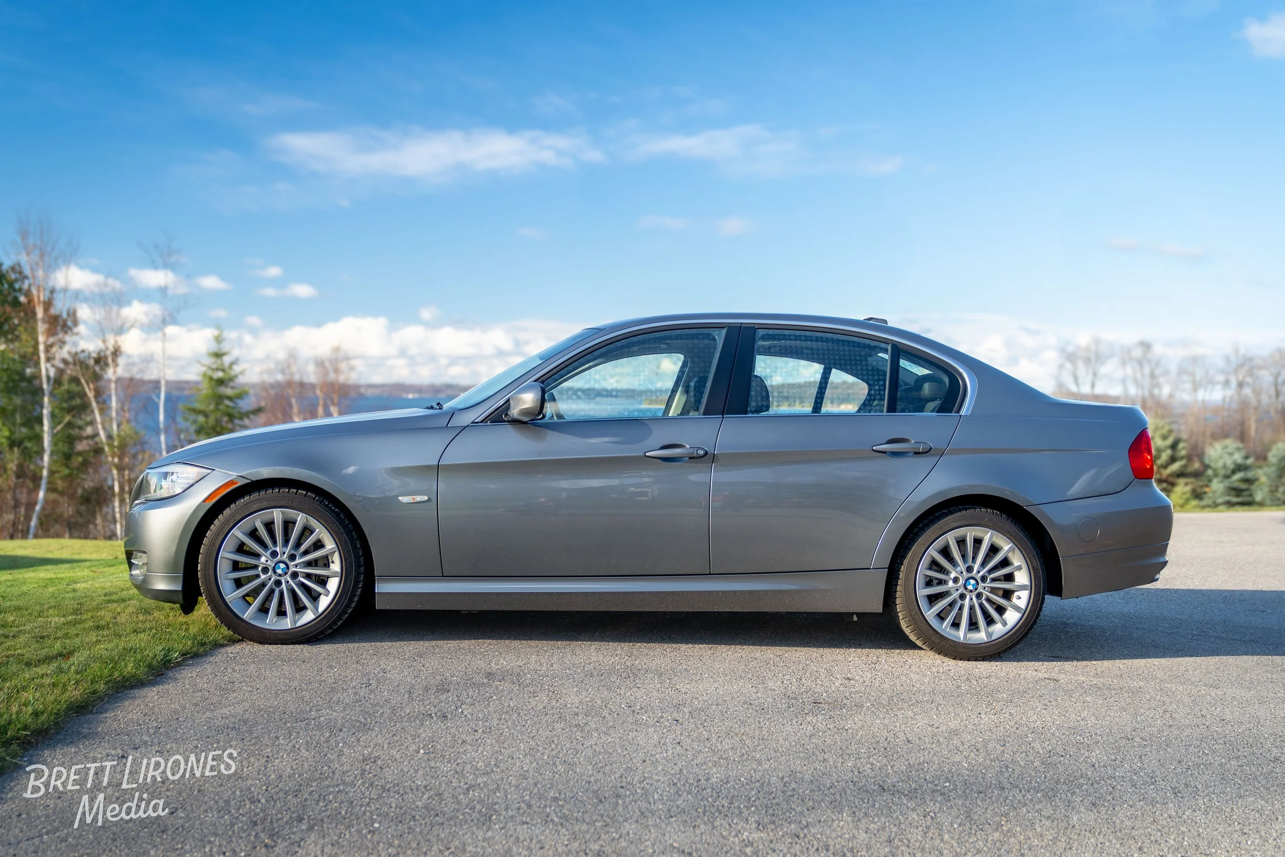 A gray BMW sedan parked on a driveway next to a grassy area, with a background of trees and a partly cloudy sky.