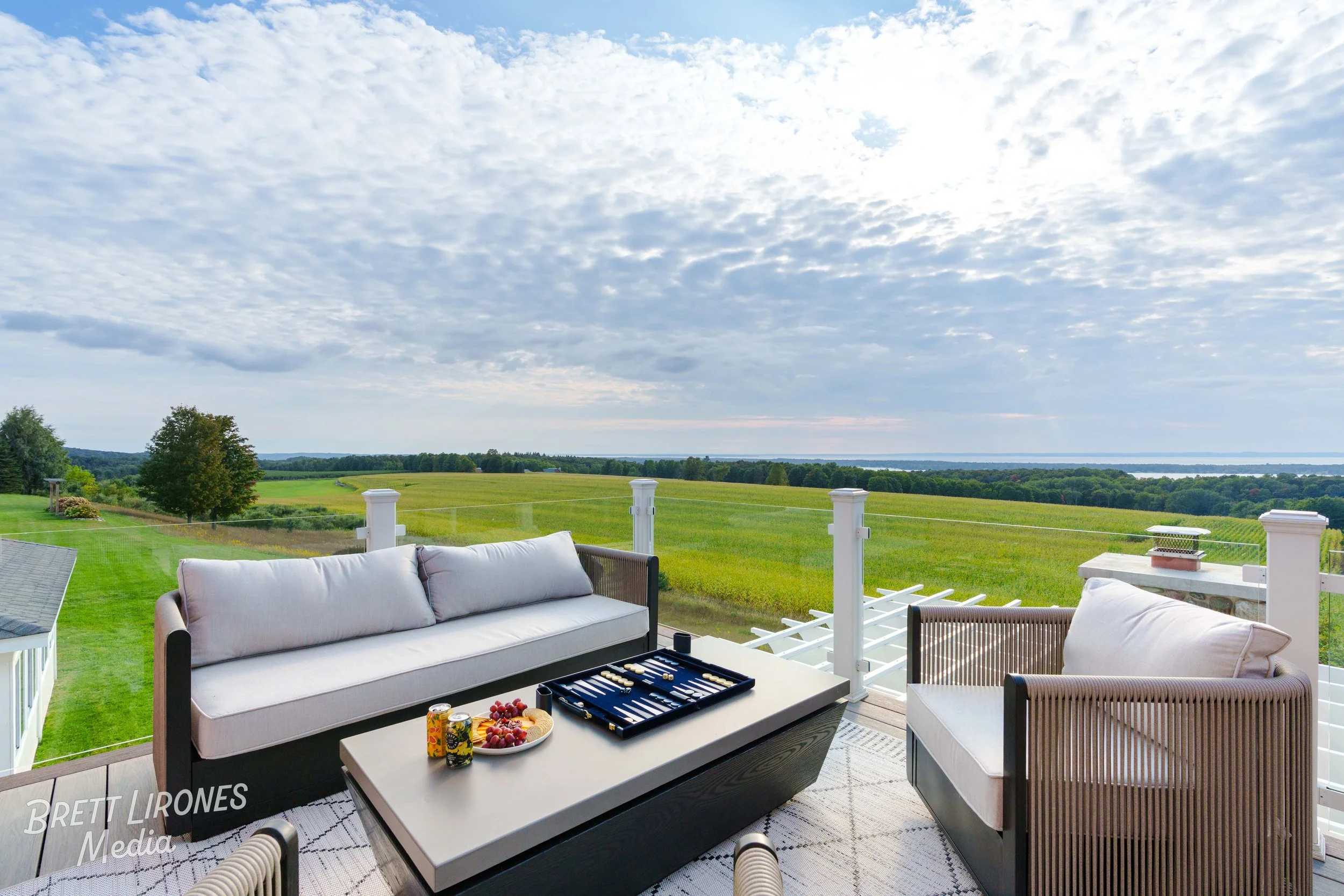 Patio with outdoor furniture including a sofa, armchair, and coffee table, overlooking open green fields and a watermark in the bottom left corner.