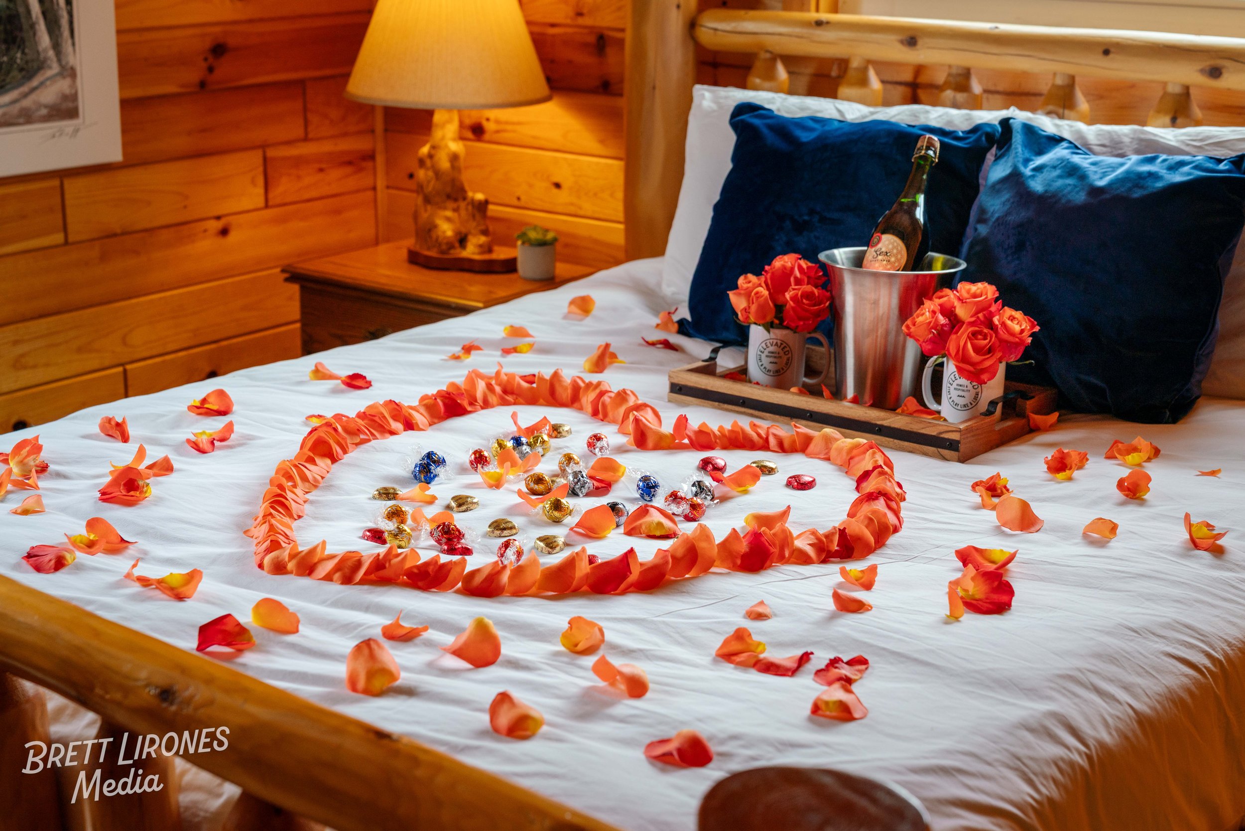A bed decorated with orange rose petals arranged in a heart shape, surrounded by chocolates and rose bouquets in mugs, with a tray holding a champagne bottle, in a cozy wooden room.