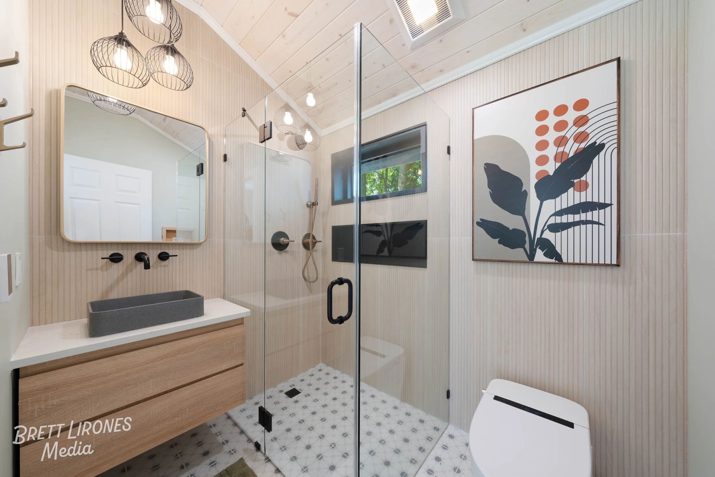 Modern bathroom with a wooden vanity, black vessel sink, rectangular mirror, glass shower enclosure, wall-mounted shower head, window with greenery outside, wall art featuring leaves, and a smart toilet.
