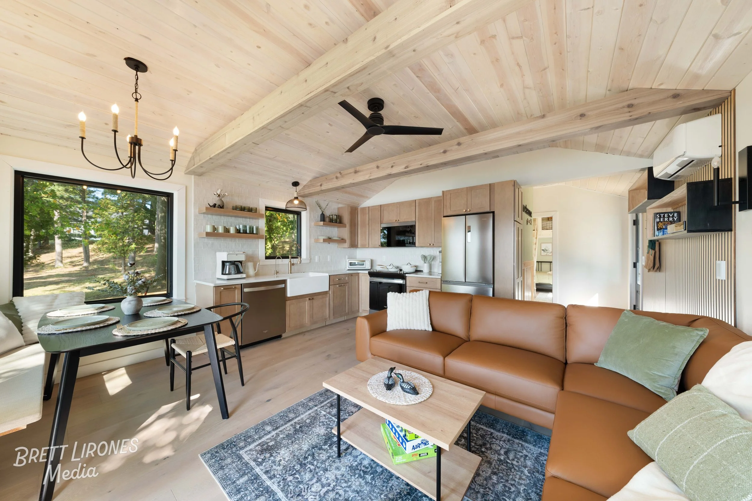 Open-concept living room and kitchen with vaulted wood ceiling, large window revealing greenery, tan leather sofa, wooden coffee table, dining table with placemats, and modern appliances.