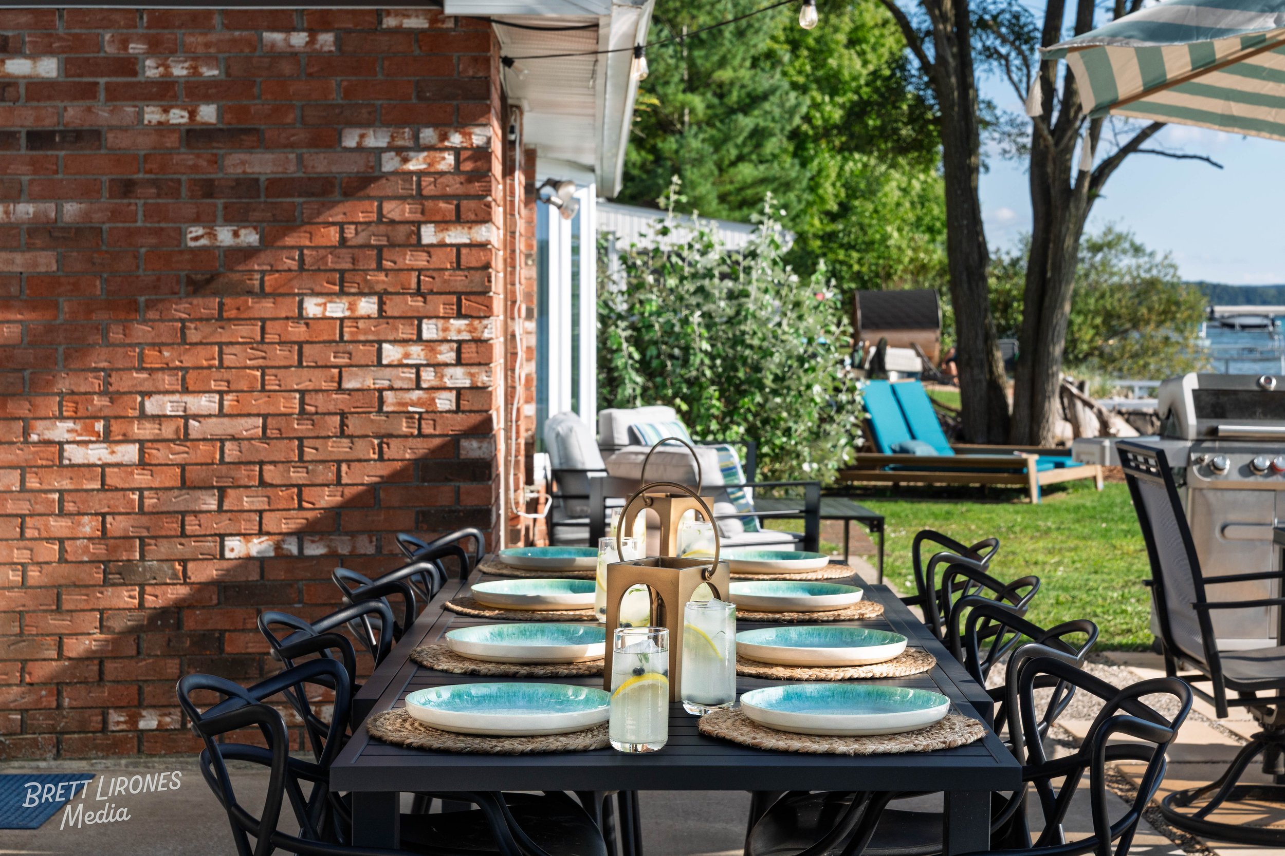 Outdoor patio dining table set for eight with plates, glasses, and a caddy with candles and lemon drinks, located beside a brick wall with greenery and a view of a lake in the background.