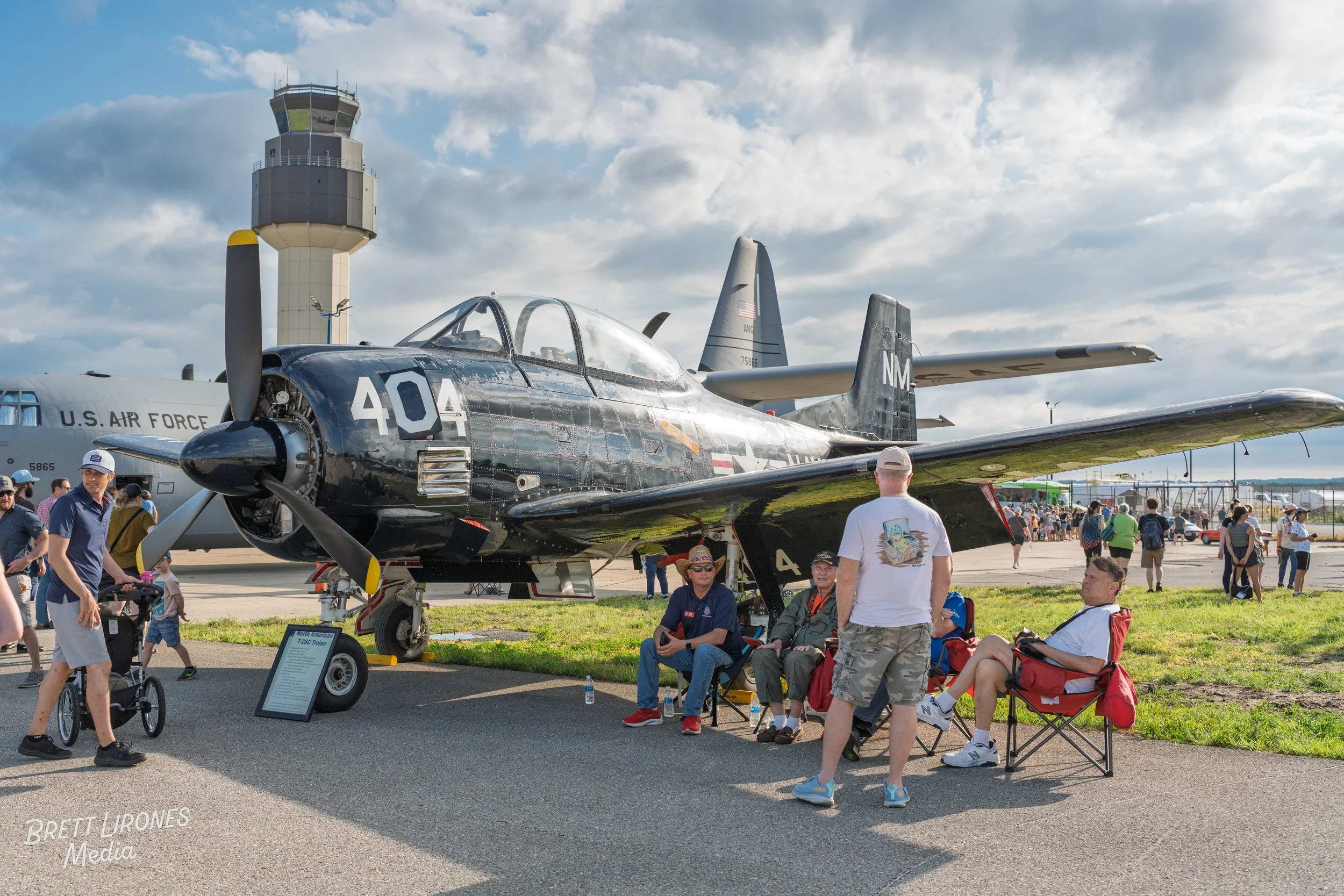 Vintage black military jet aircraft displayed outdoors at an airshow, with a control tower and people walking and sitting nearby.