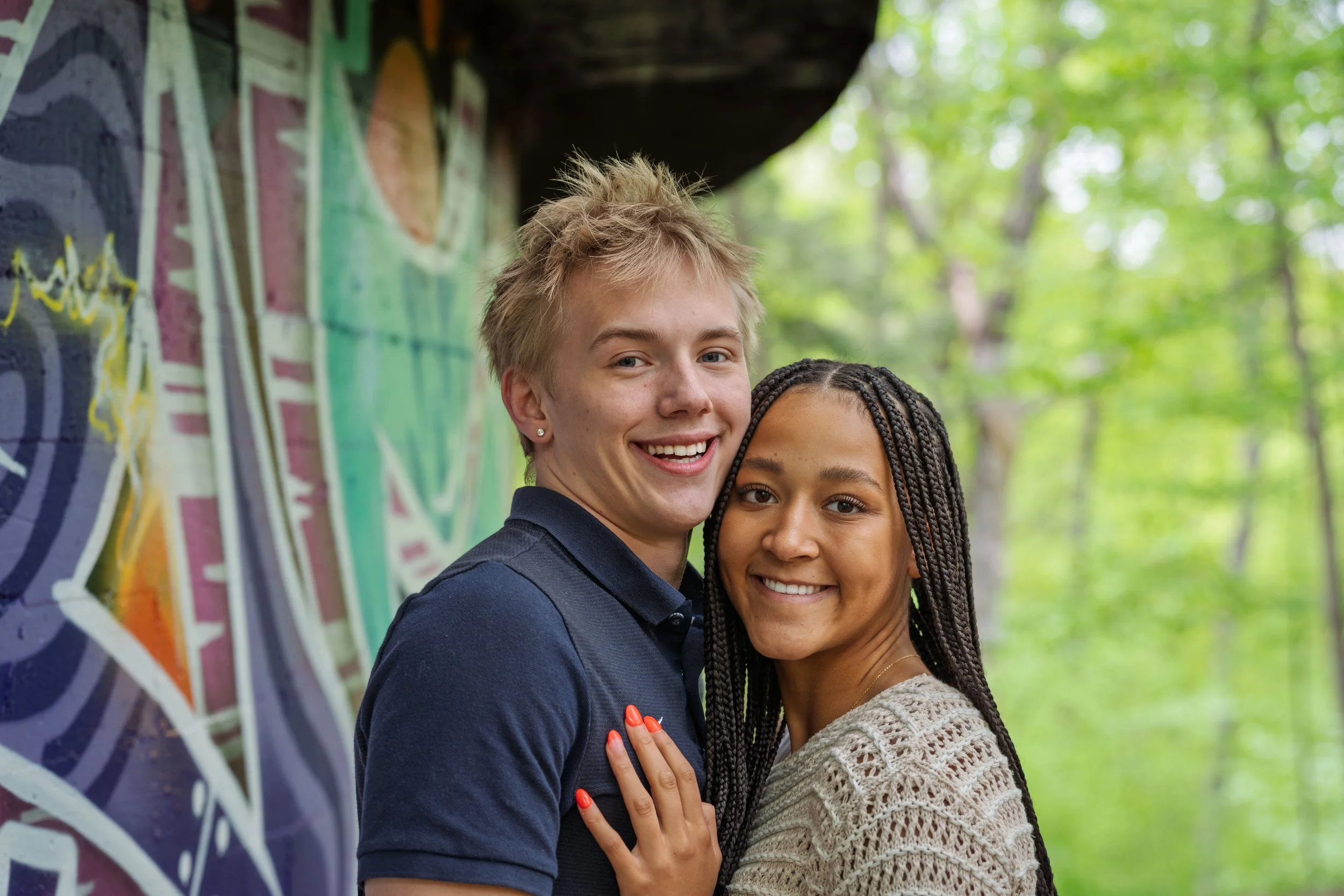 A smiling young couple embracing outdoors with a graffiti-covered wall and lush green trees in the background.