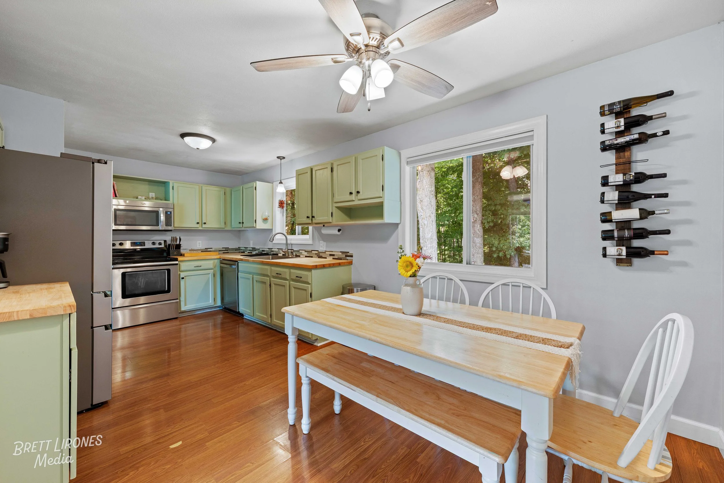 Kitchen with light green cabinets, stainless steel appliances, a white dining table with four chairs, a window showing trees outside, and a ceiling fan with light fixtures.