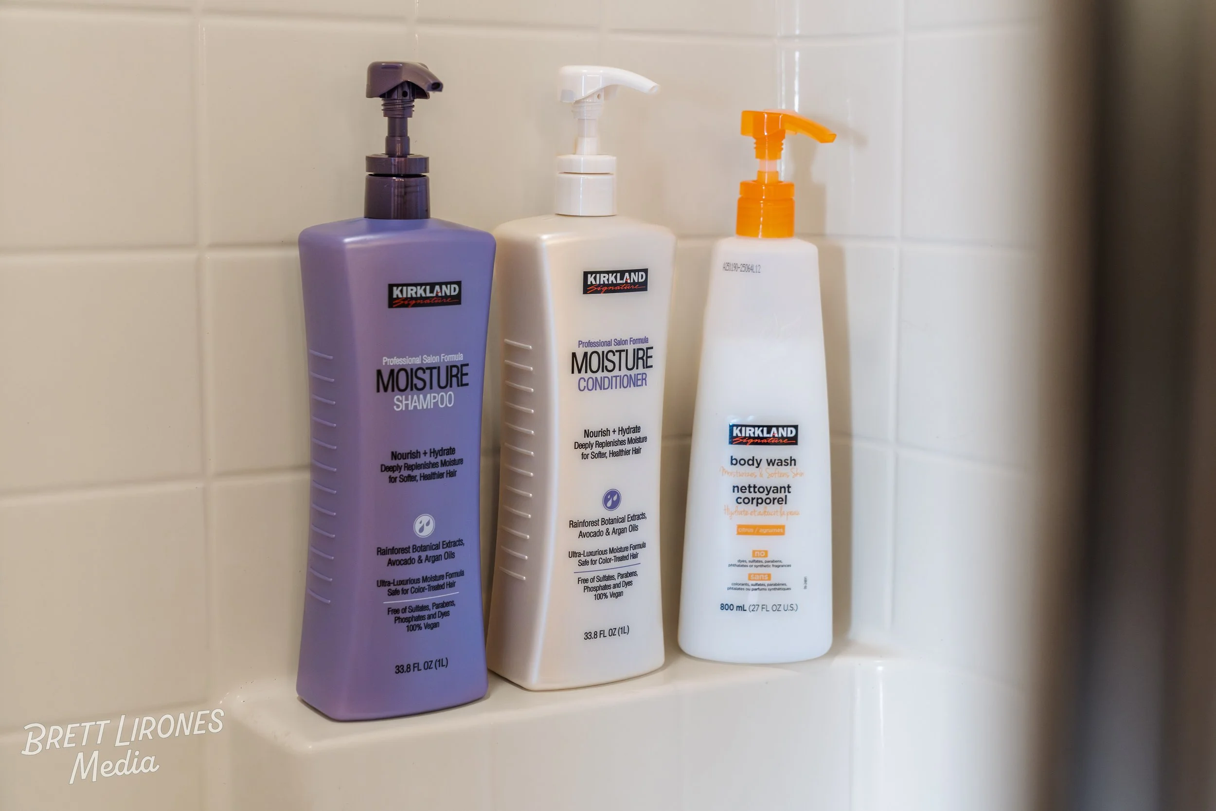 Three Kirkland personal care bottles in a shower corner: moisturizing shampoo, moisturizing conditioner, and body wash, against a white tiled wall.