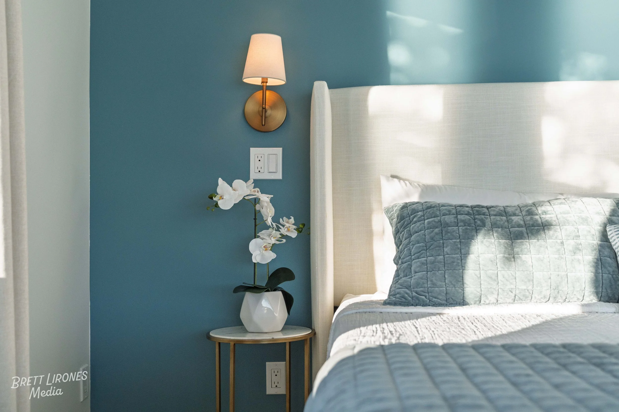 A bedroom corner with a beige headboard, quilted pillow, nightstand, orchid plant, blue accent wall, wall-mounted lamp, and electrical outlets.
