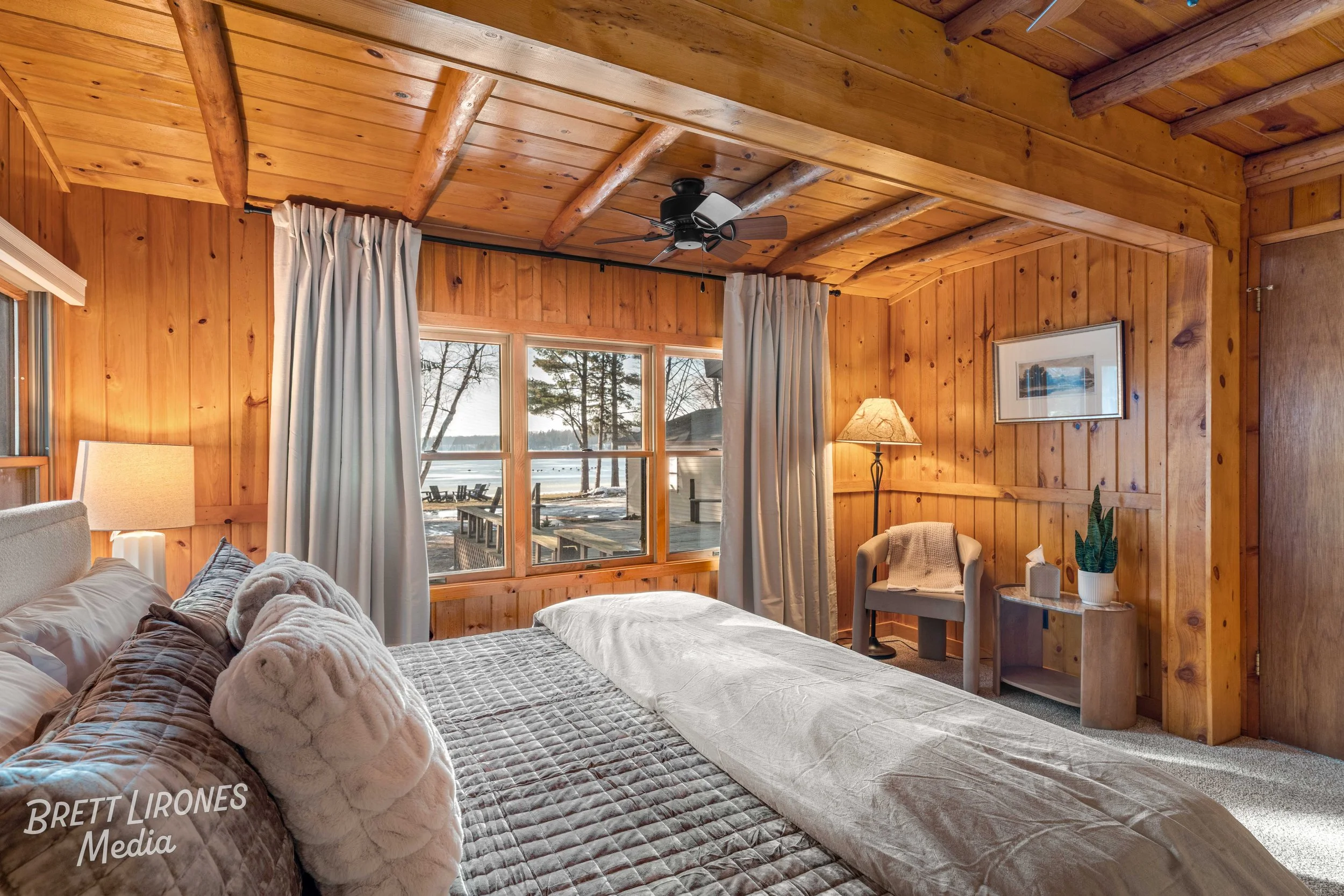 Cozy bedroom with wooden panel walls and ceiling, large window with curtains, bedside lamp, a framed picture, comfortable bed with plush pillows and rolled towels, and outside view of a lake and trees.