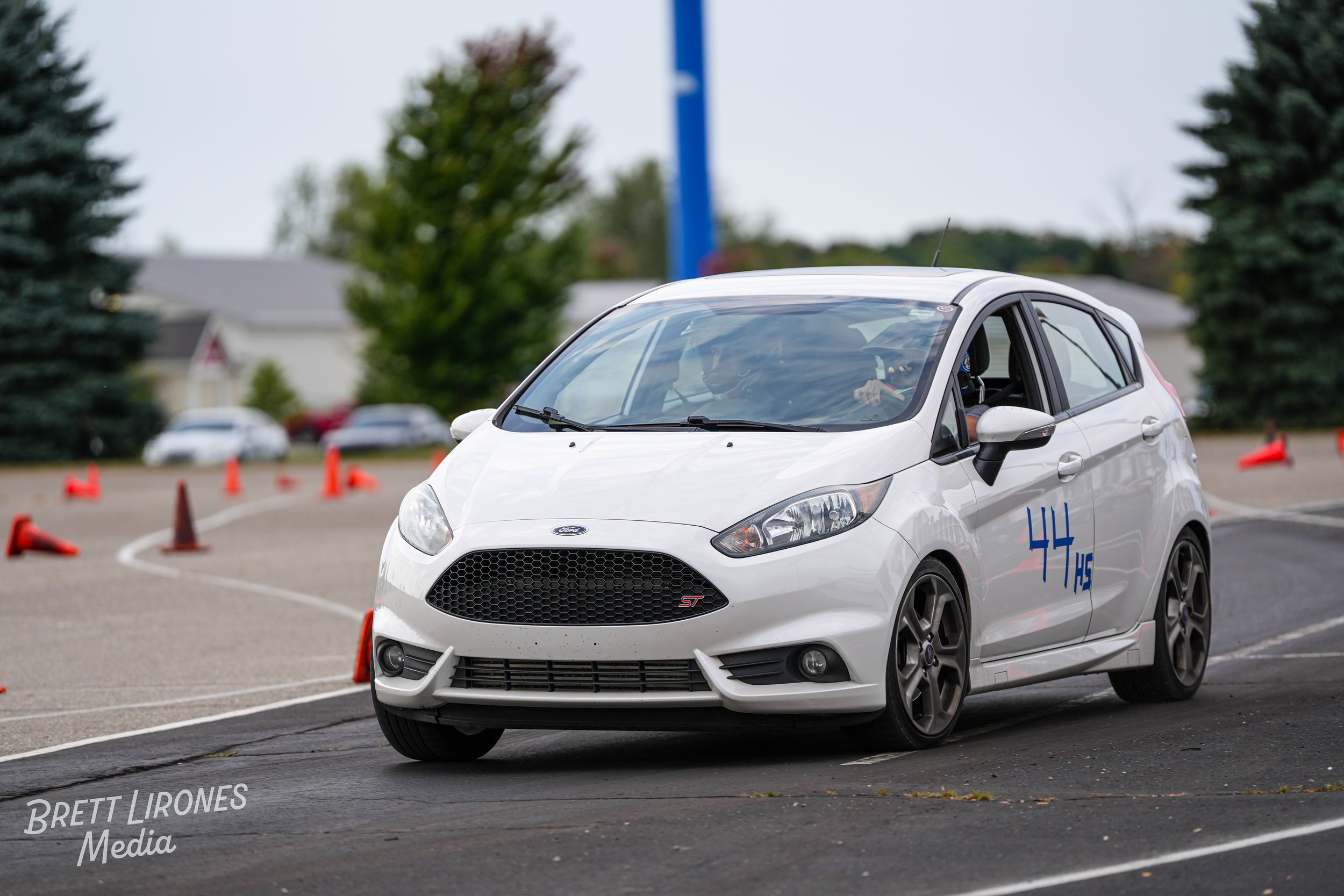 A white Ford Fiesta ST hatchback participating in a driving course, navigating around orange traffic cones on an asphalt track, with a driver wearing a helmet inside.