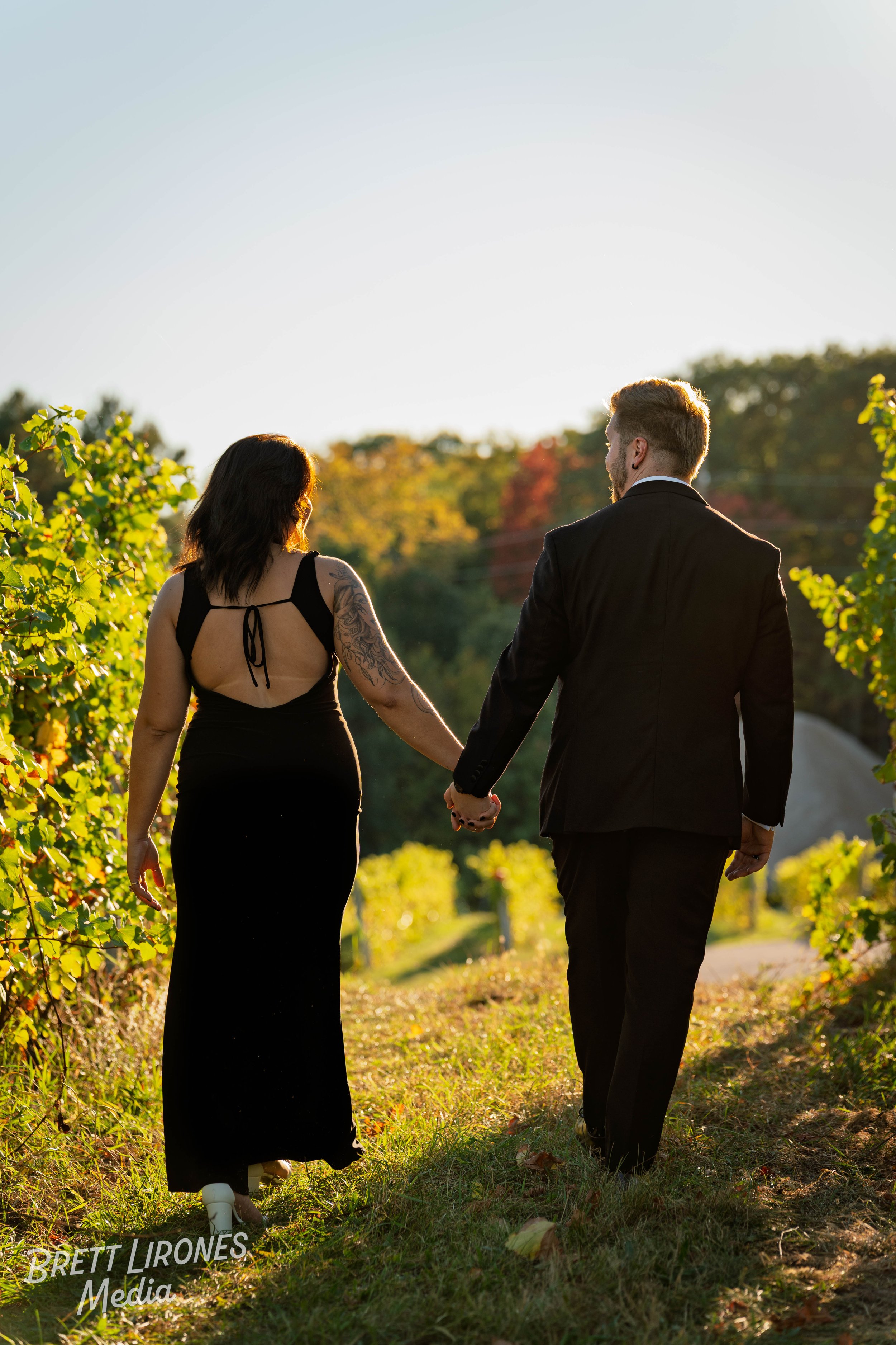 A couple holding hands and walking through a vineyard during sunset, with autumn-colored trees in the background.