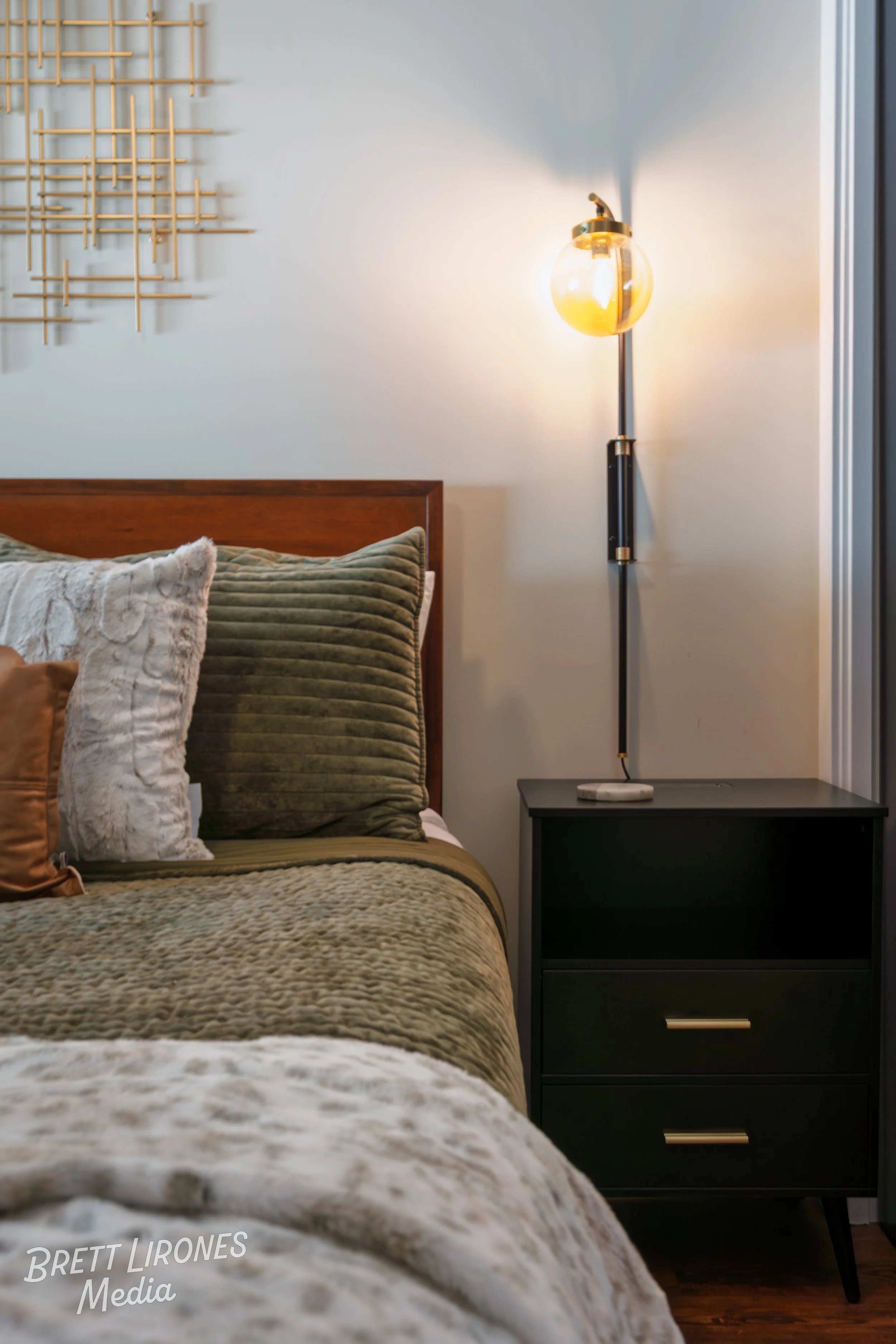 A bedroom with a bed featuring plush pillows and a beige blanket, a black nightstand with gold handles, a modern floor lamp with a round yellow bulb, and wall art above the bed.