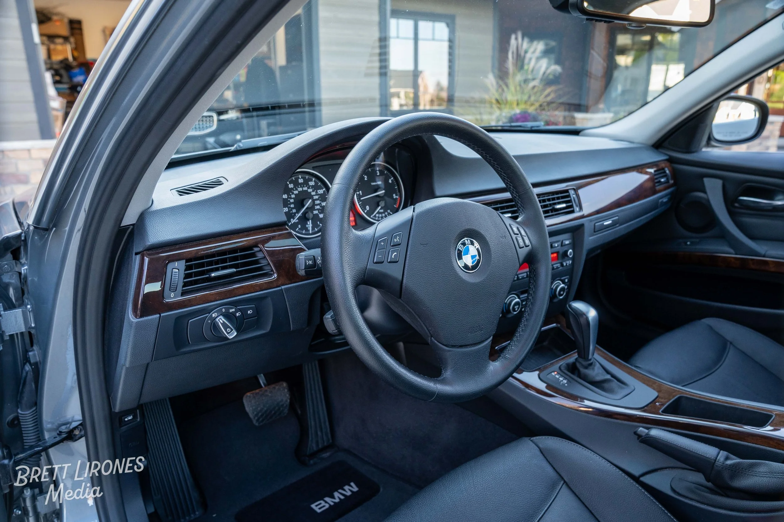 Interior of a BMW car showing the steering wheel with BMW logo, dashboard with speedometer and tachometer, gear shifter, and wood paneling on the dashboard and center console.