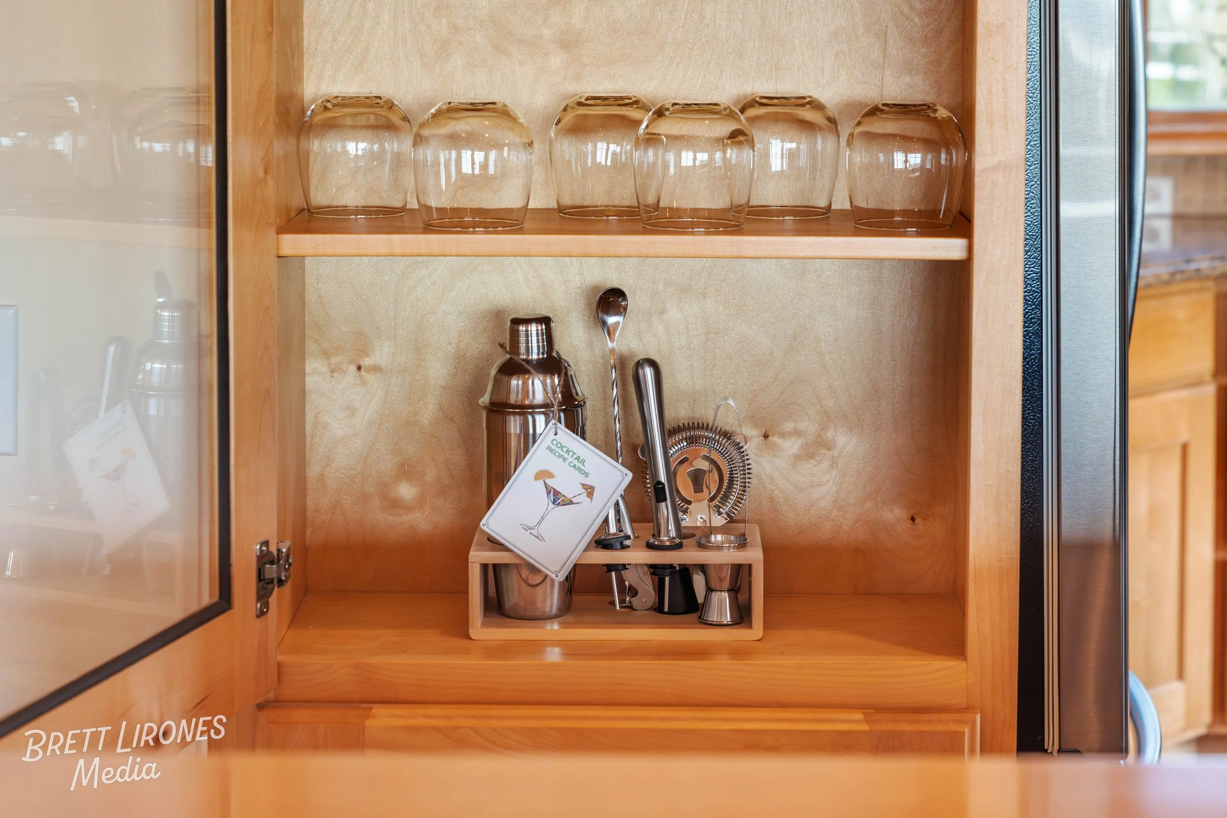 Open wooden cabinet with empty glass cups on top shelf and bar tools on the bottom shelf, including a cocktail shaker with a drink recipe card attached, a spoon, a strainer, a muddler, and a jigger.