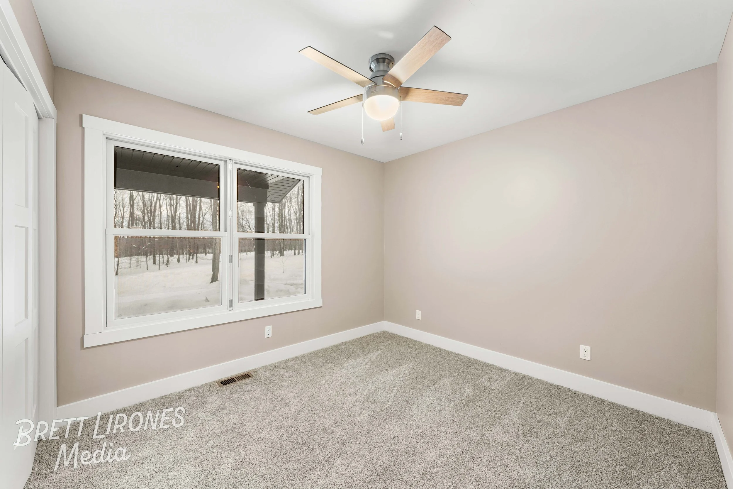 Empty bedroom with beige walls, a large window showing a snowy landscape, a ceiling fan, and carpeted floor.