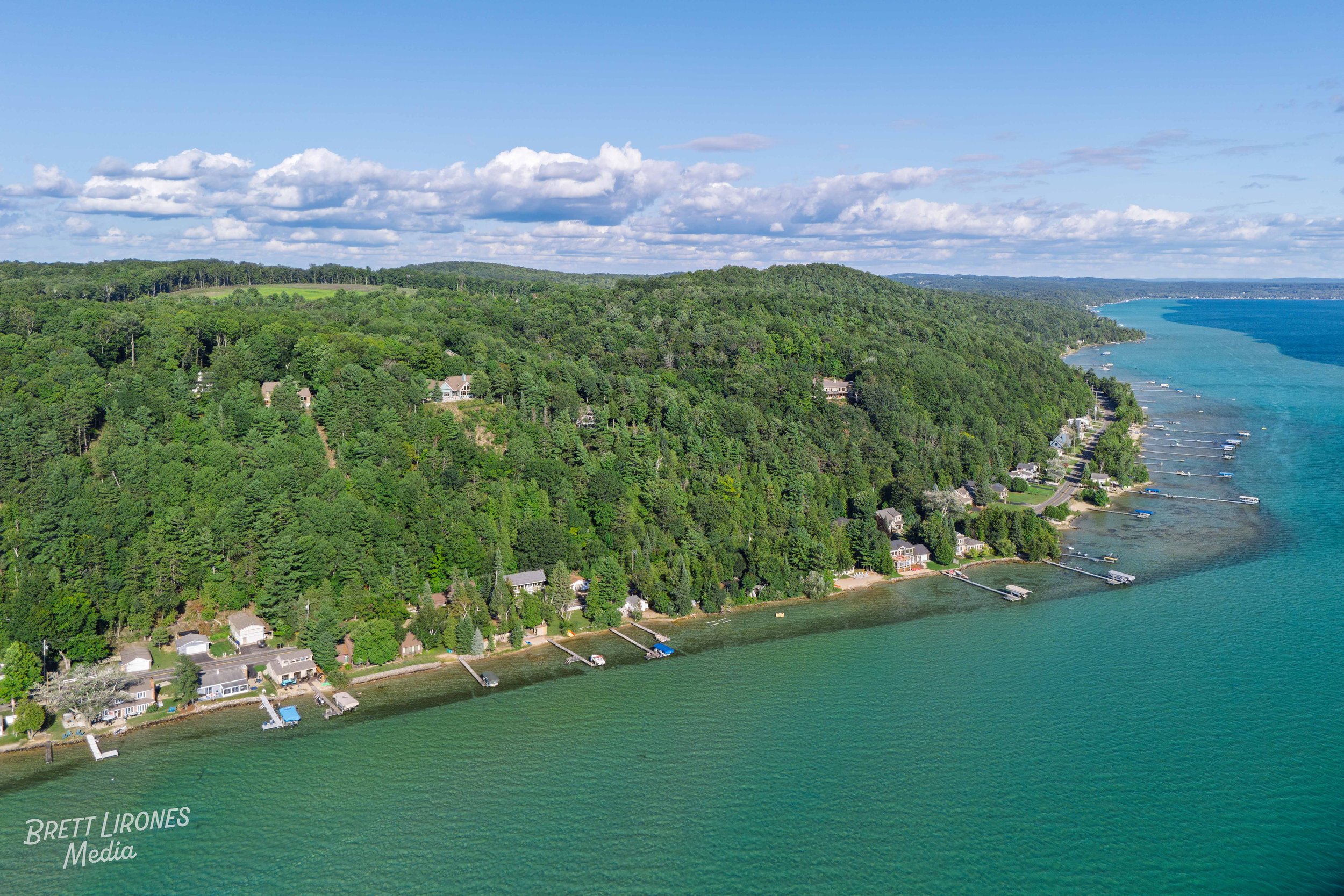 Aerial view of a shoreline with houses and docks along a green, forested hillside next to blue-green water, under a partly cloudy sky.