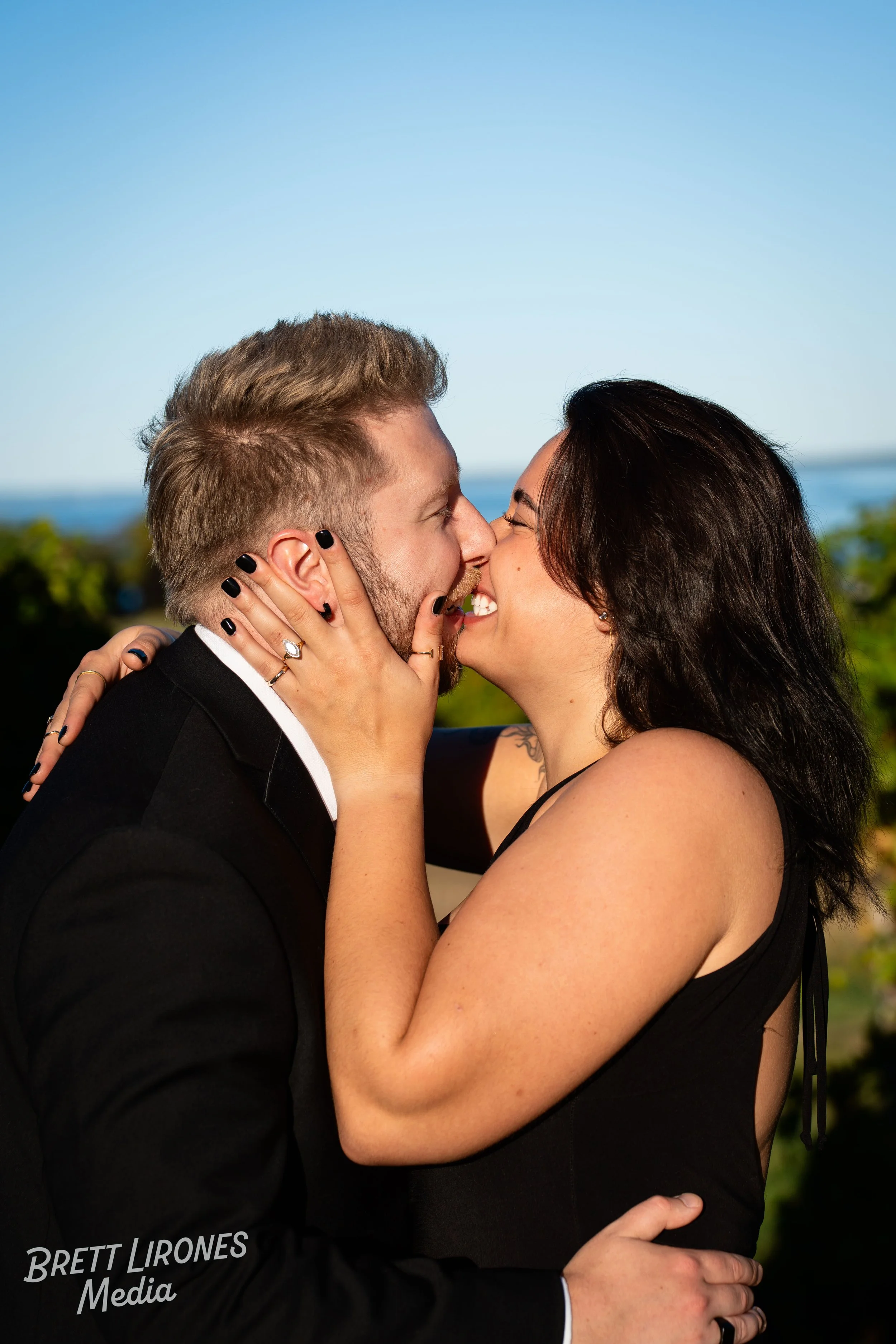 A couple passionately kissing outdoors, with the man dressed in a wedding suit and the woman in a black dress.
