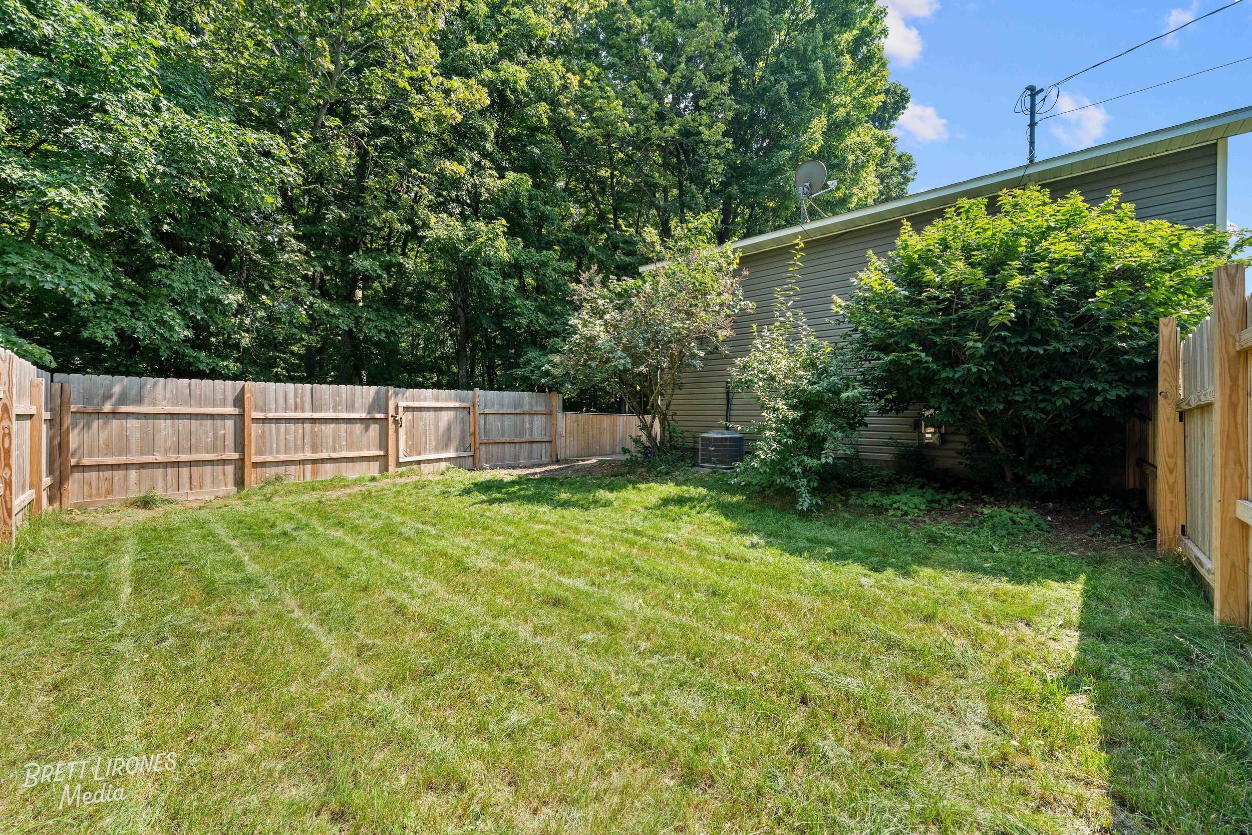 A fenced backyard with green grass, shrubs, trees, and a house with satellite dishes and an air conditioning unit.