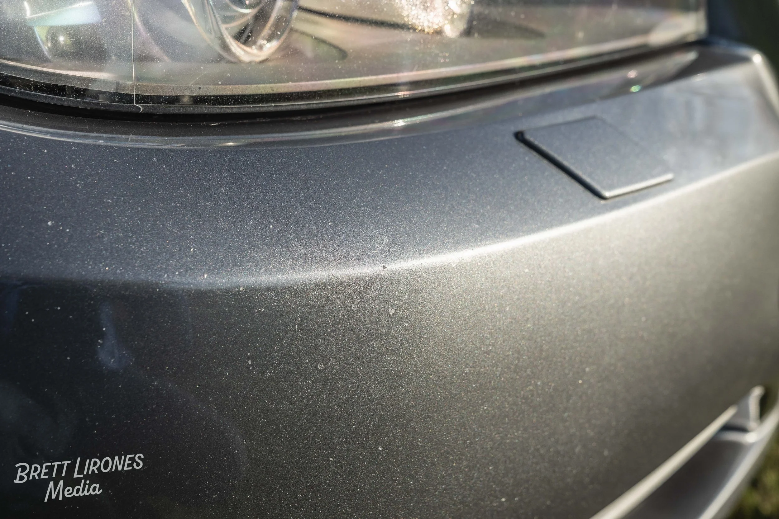 Close-up of a gray car's front bumper and headlight with some dust and small scratches.