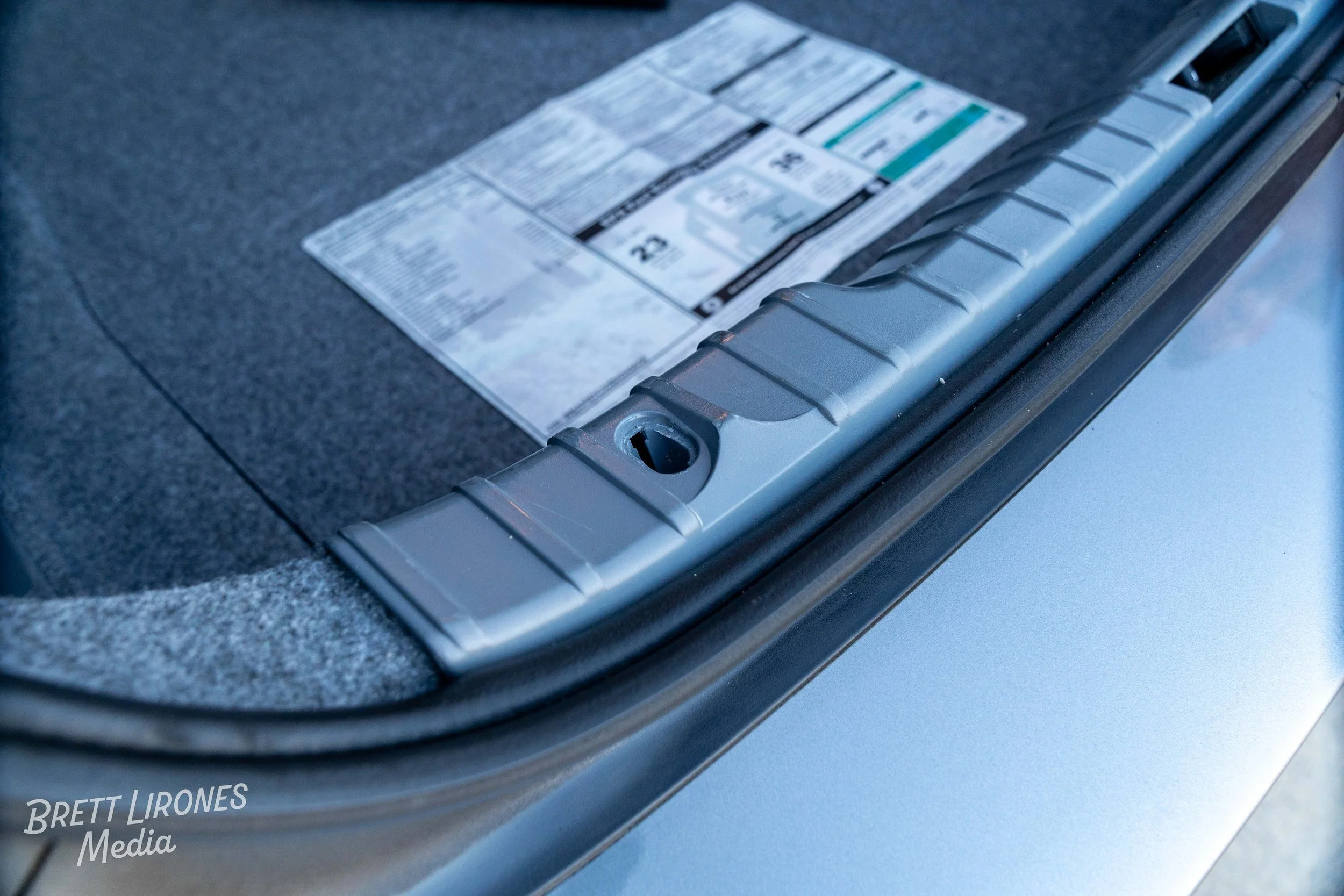 Close-up of a car trunk with an empty lock hole, carpeted interior, and a transparent sticker with information on the trunk floor.