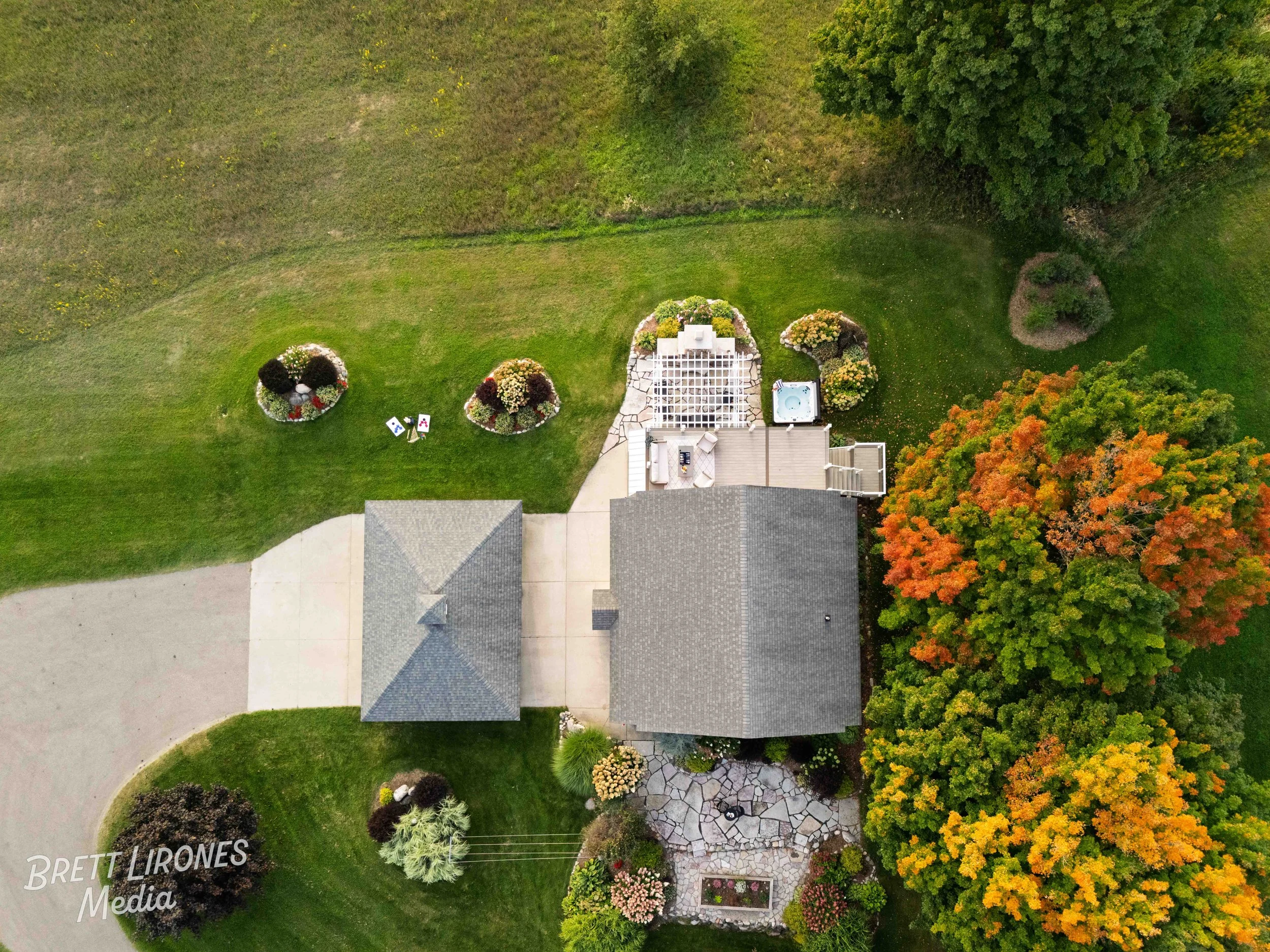 An aerial view of a house with a gray roof, surrounded by colorful autumn trees, a green lawn, and a landscaped backyard with a hot tub, patio furniture, and decorative bushes.