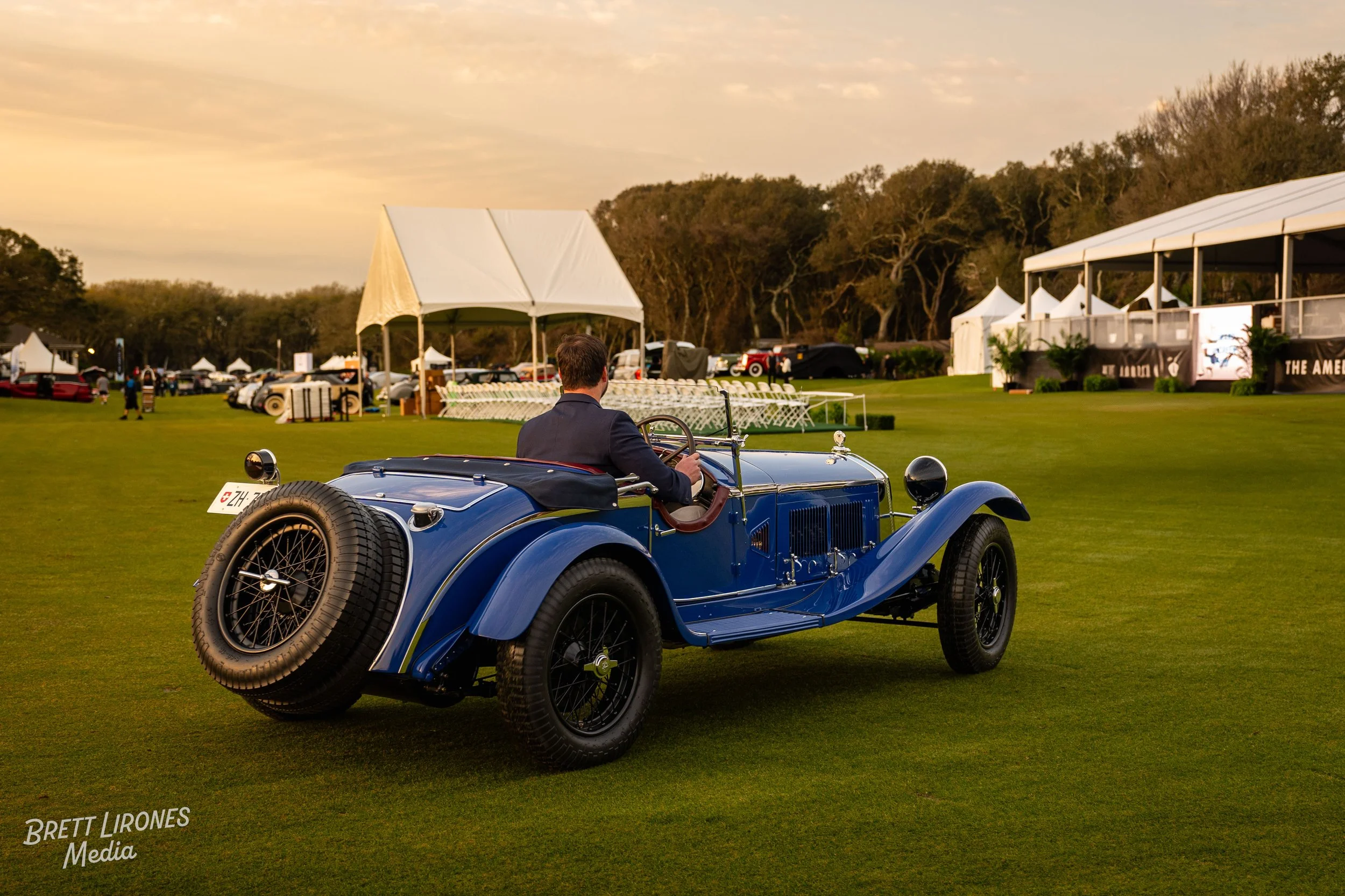 A vintage blue convertible car with a person in formal attire sitting in the driver's seat on a lush green lawn at a classic car event during sunset.
