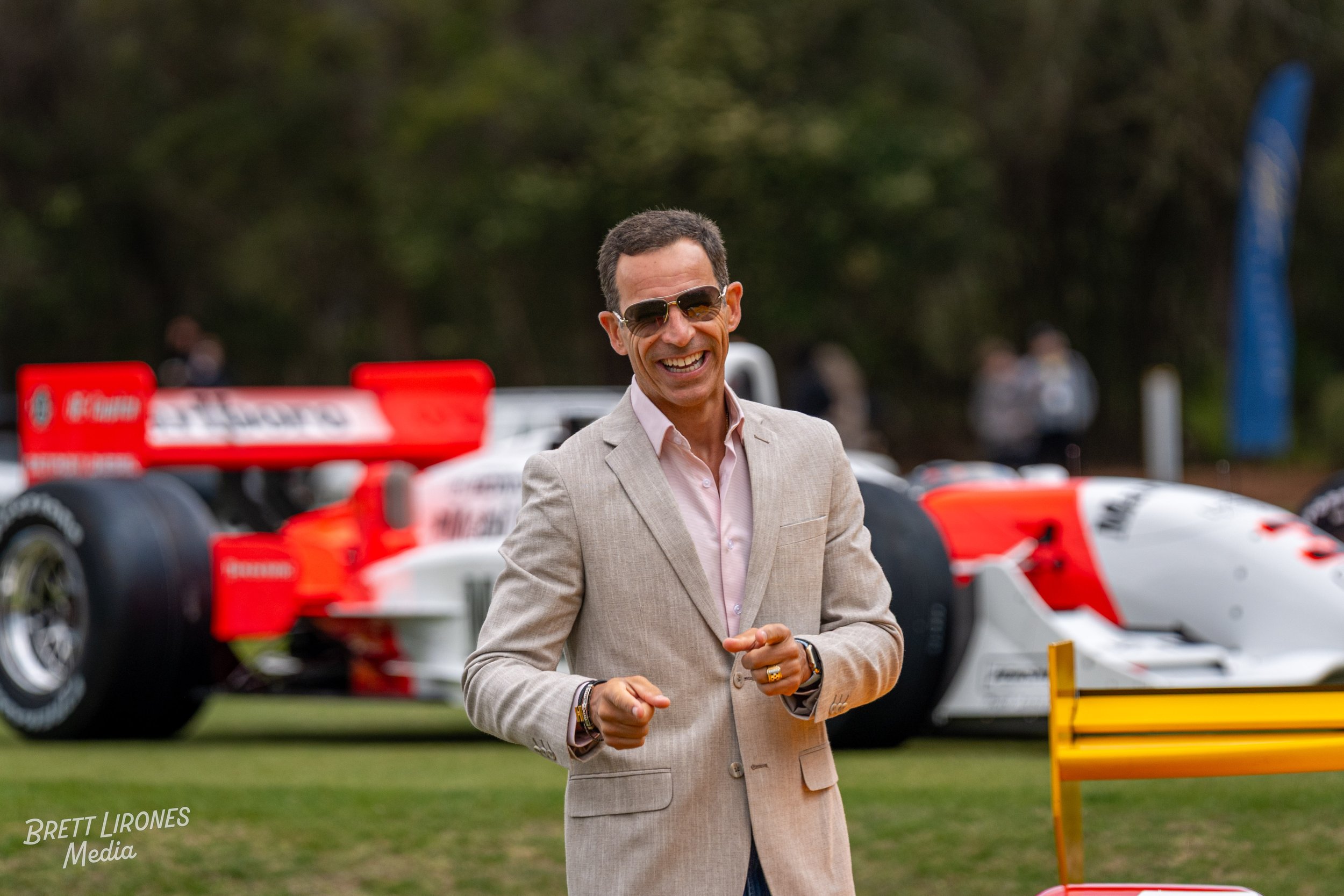 Smiling man wearing sunglasses and a beige suit standing outdoors with race cars in the background.