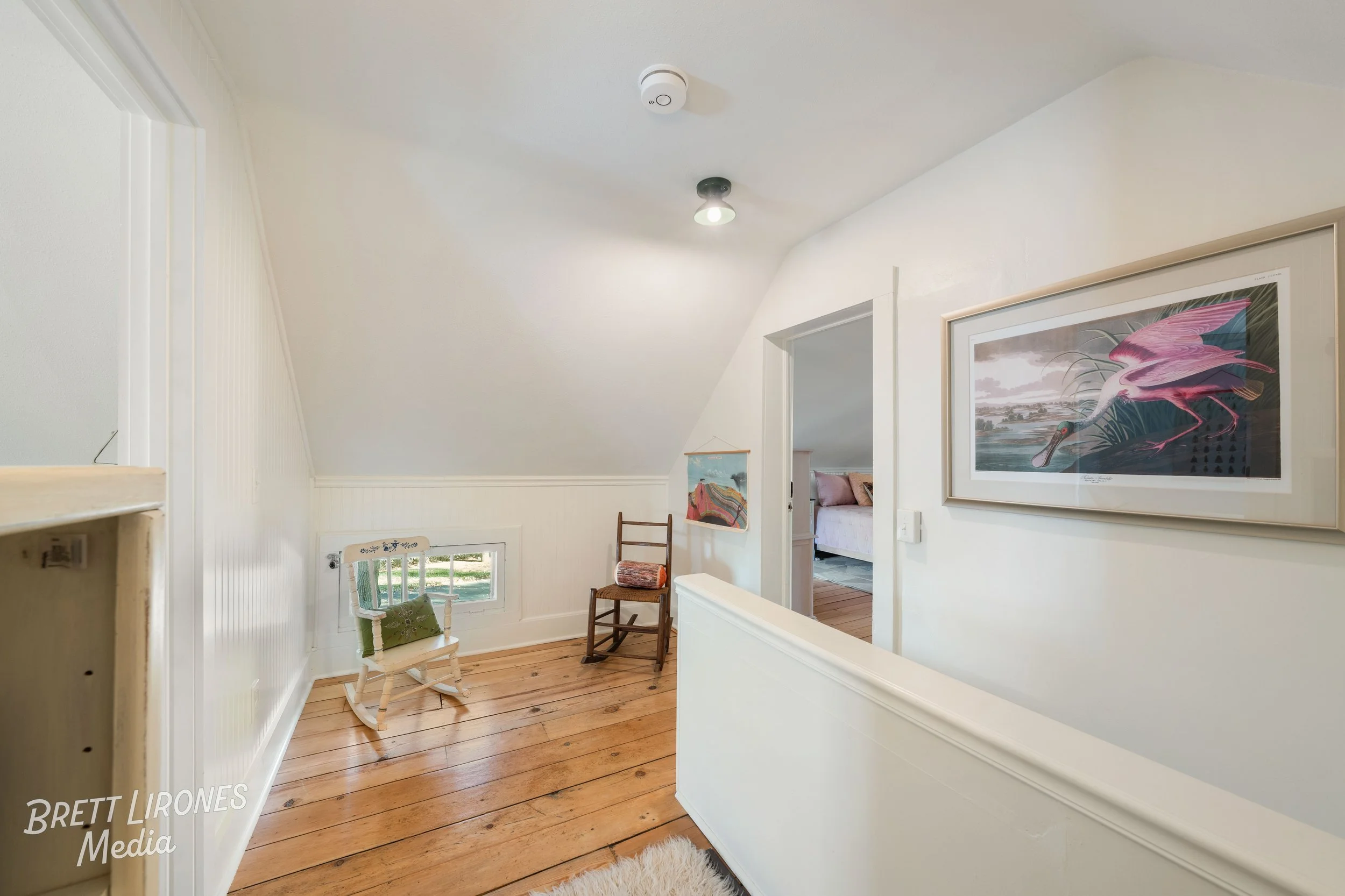 A small landing area with wood flooring, white walls, a framed pink bird artwork, a small window, a white wooden rocking chair with a green pillow, and a brown wooden rocking chair. Part of a bedroom with a white bed and pink pillows is visible throu