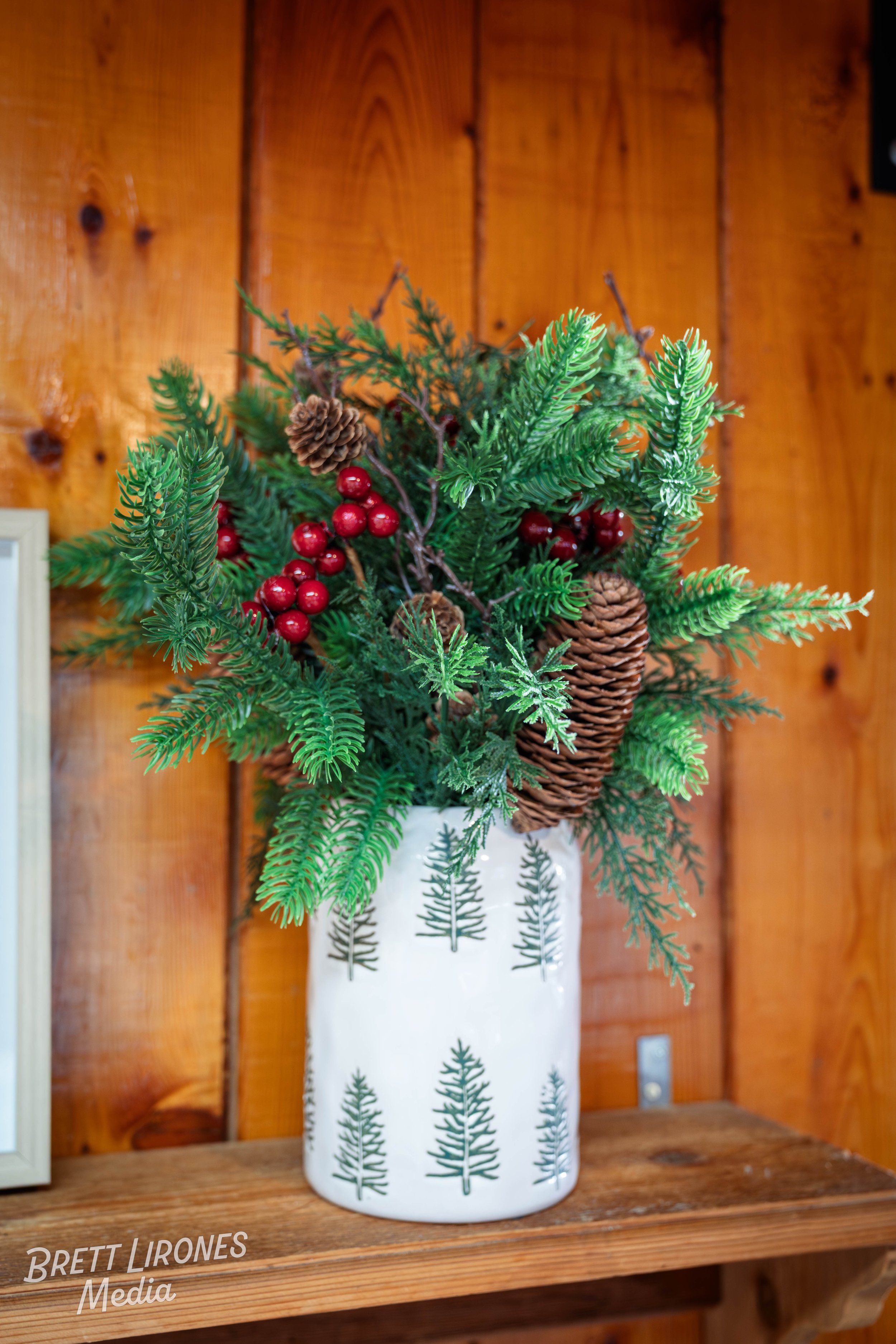 A Christmas-themed floral arrangement in a white vase decorated with green evergreen tree patterns, featuring pinecones, red berries, and various green foliage, set against a wooden background.