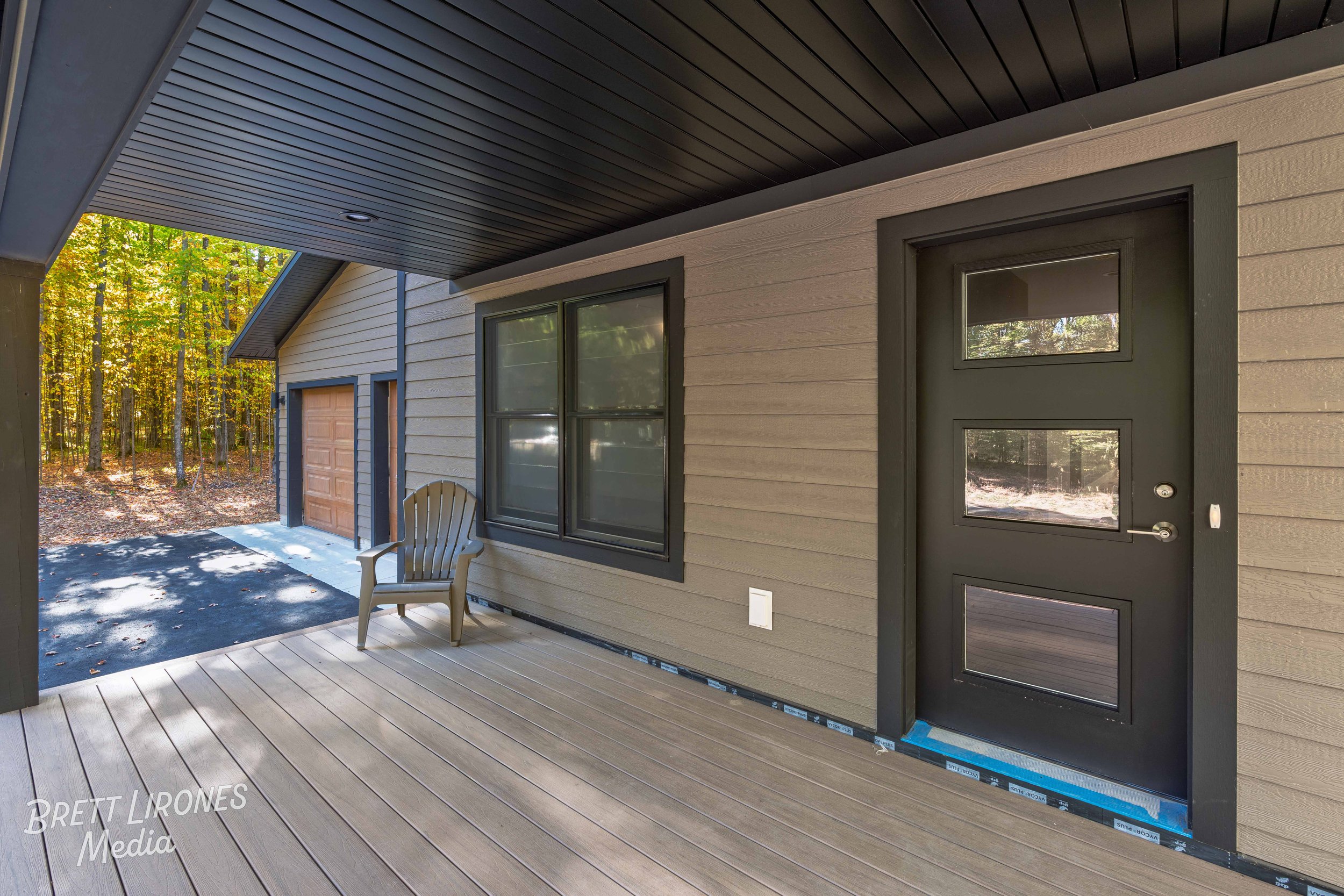 Modern house porch with a black-framed door with three glass panels, beige siding, a window with black trim, a grey Adirondack chair, and a wooded yard with fall foliage in the background.
