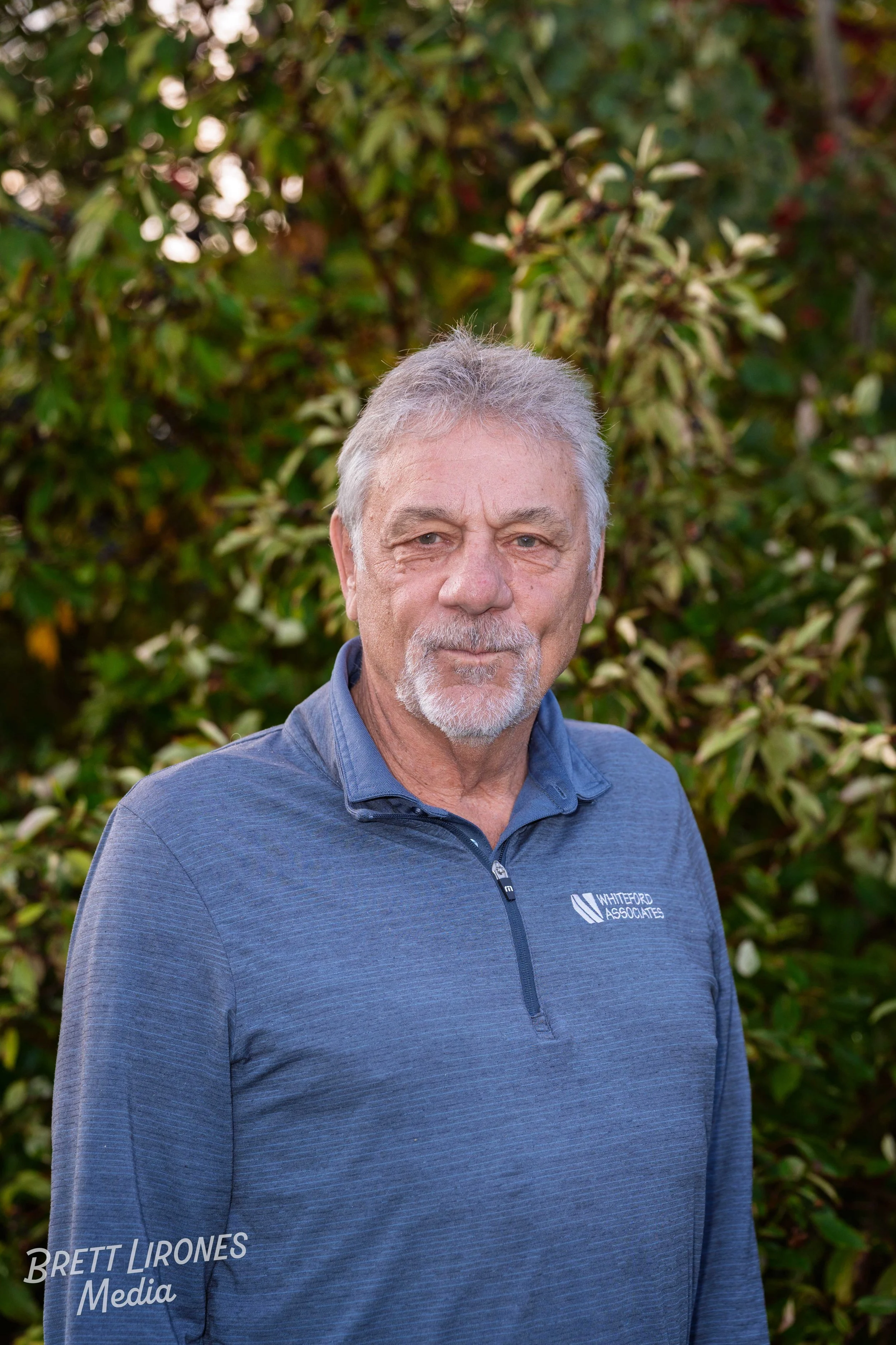 A middle-aged man with gray hair and a beard standing outdoors in front of green and brown foliage, wearing a blue quarter-zip pullover with a logo.