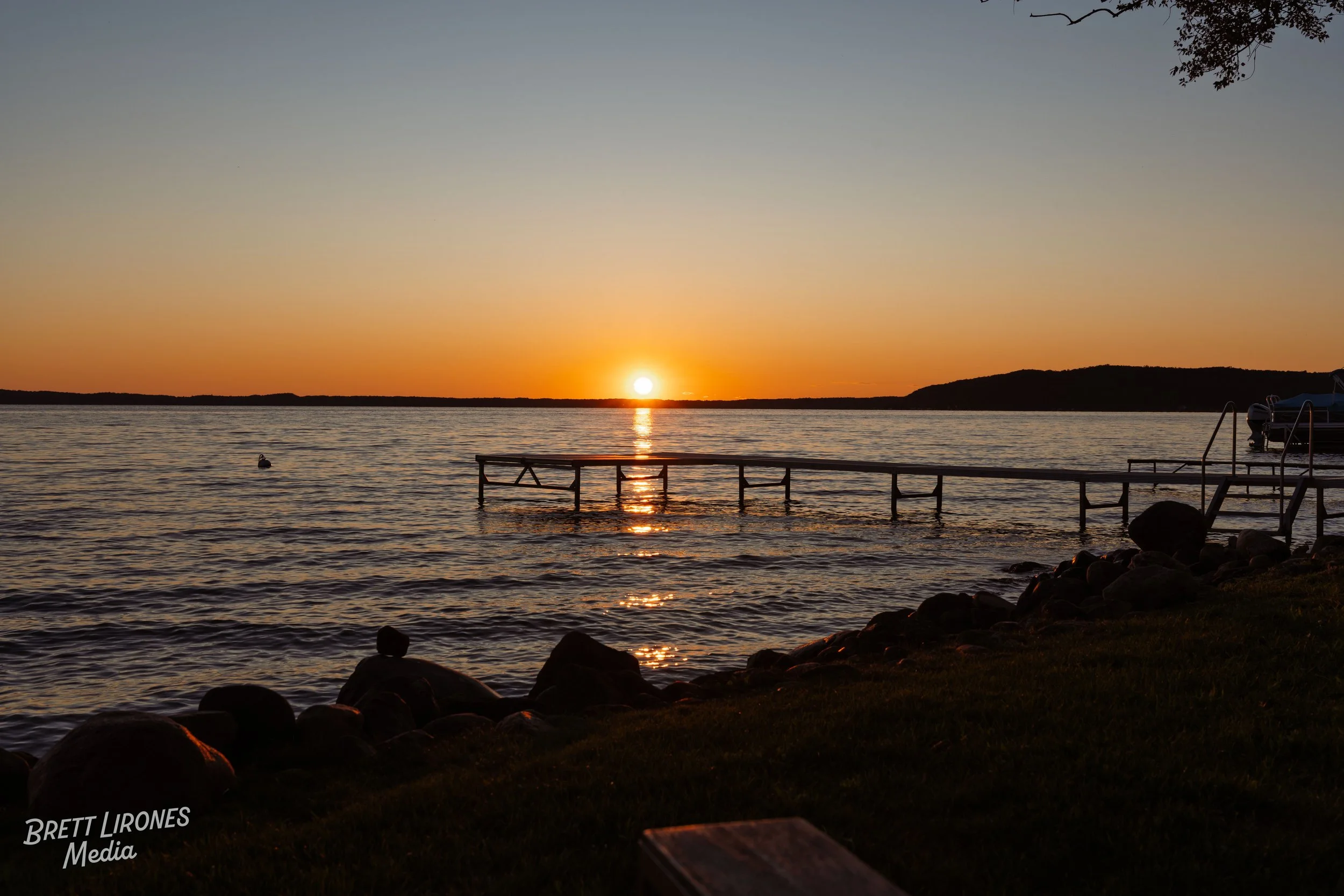 Sunset over a lake with a wooden dock extending into the water, surrounded by rocks and grassy shoreline, with trees and hills in the background.