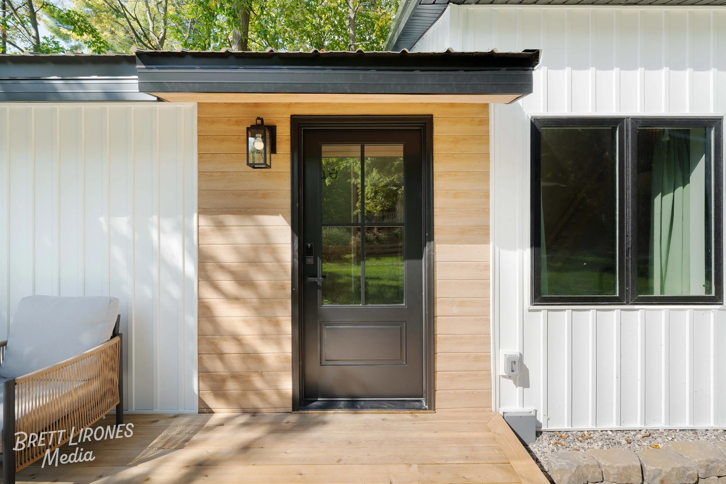 Front view of a modern house with wooden and white siding, black door with glass panels, a window with black framing, an outdoor chair to the left, and a small outdoor light fixture.