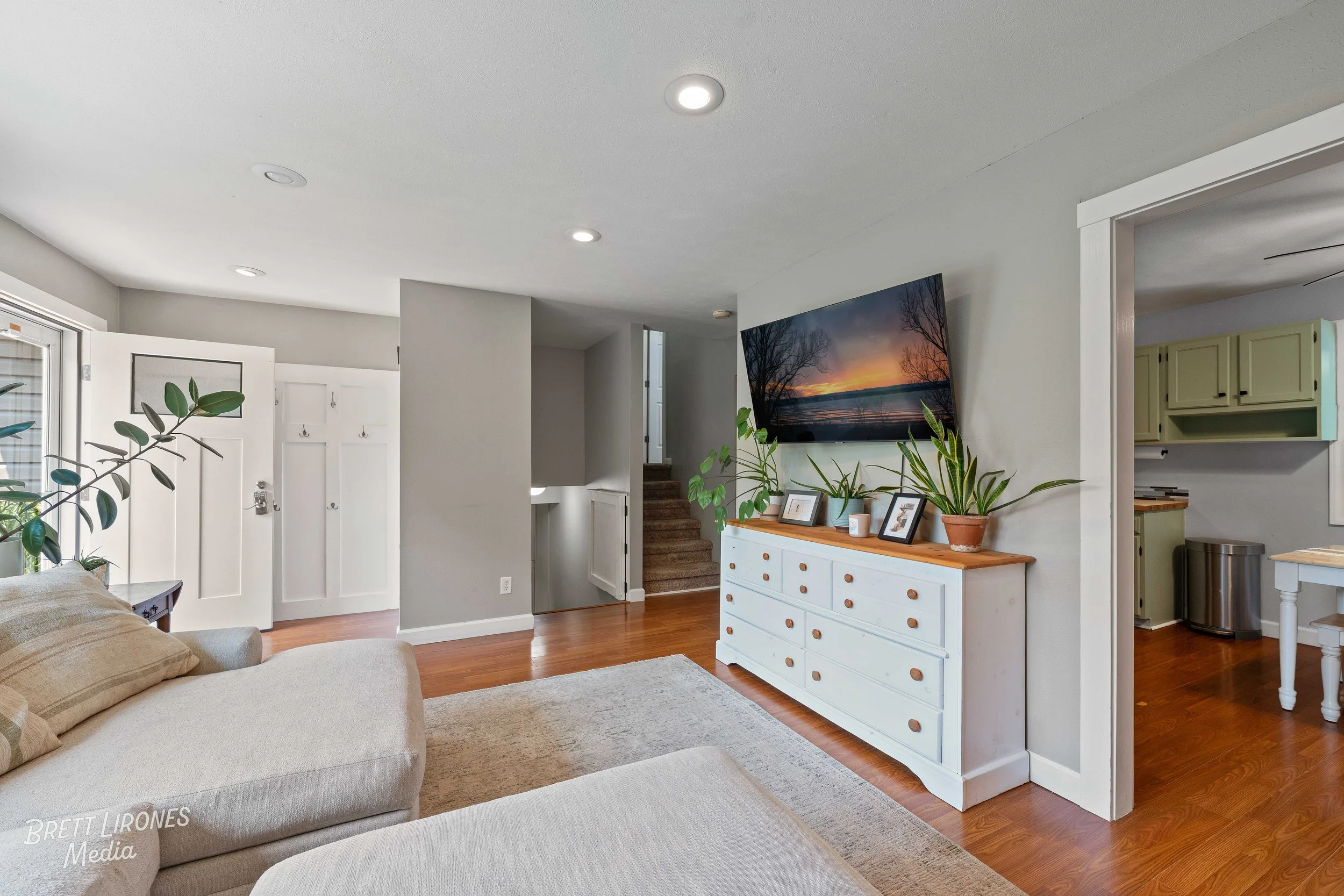 Living room with beige sofa, white dresser with plants and picture frames, wall-mounted TV, hardwood floors, and kitchen partially visible through doorway.