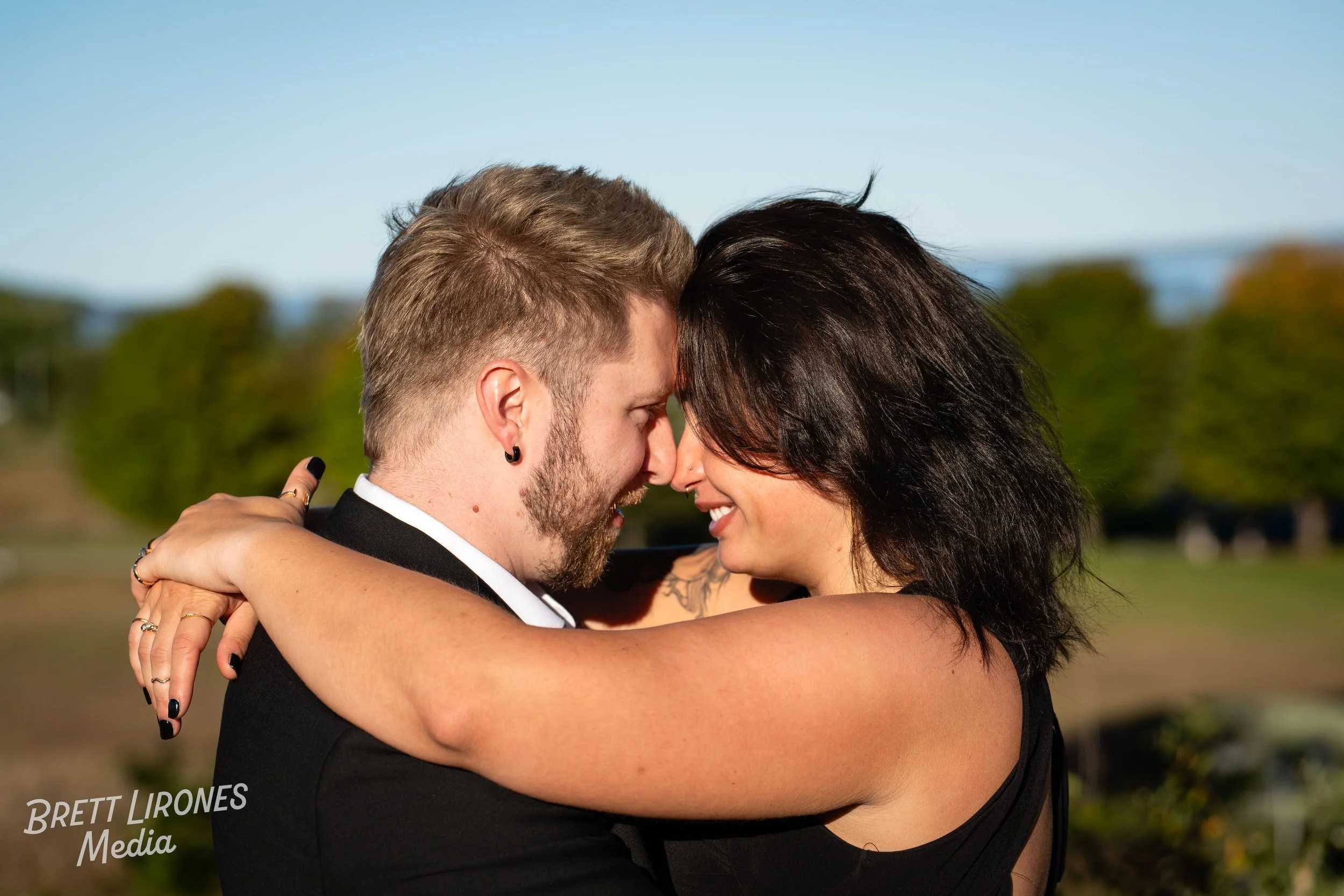 A couple embracing outdoors with their faces close together, smiling, and arms wrapped around each other, during daytime.