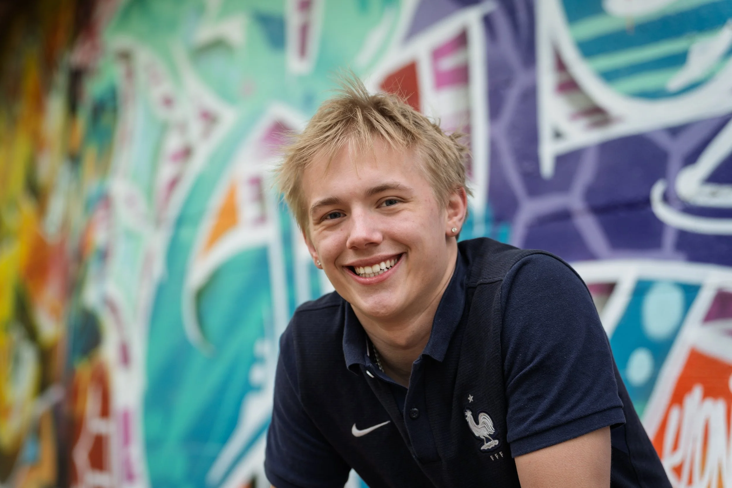 A young man with blonde hair and earrings smiling in front of a colorful graffiti wall.