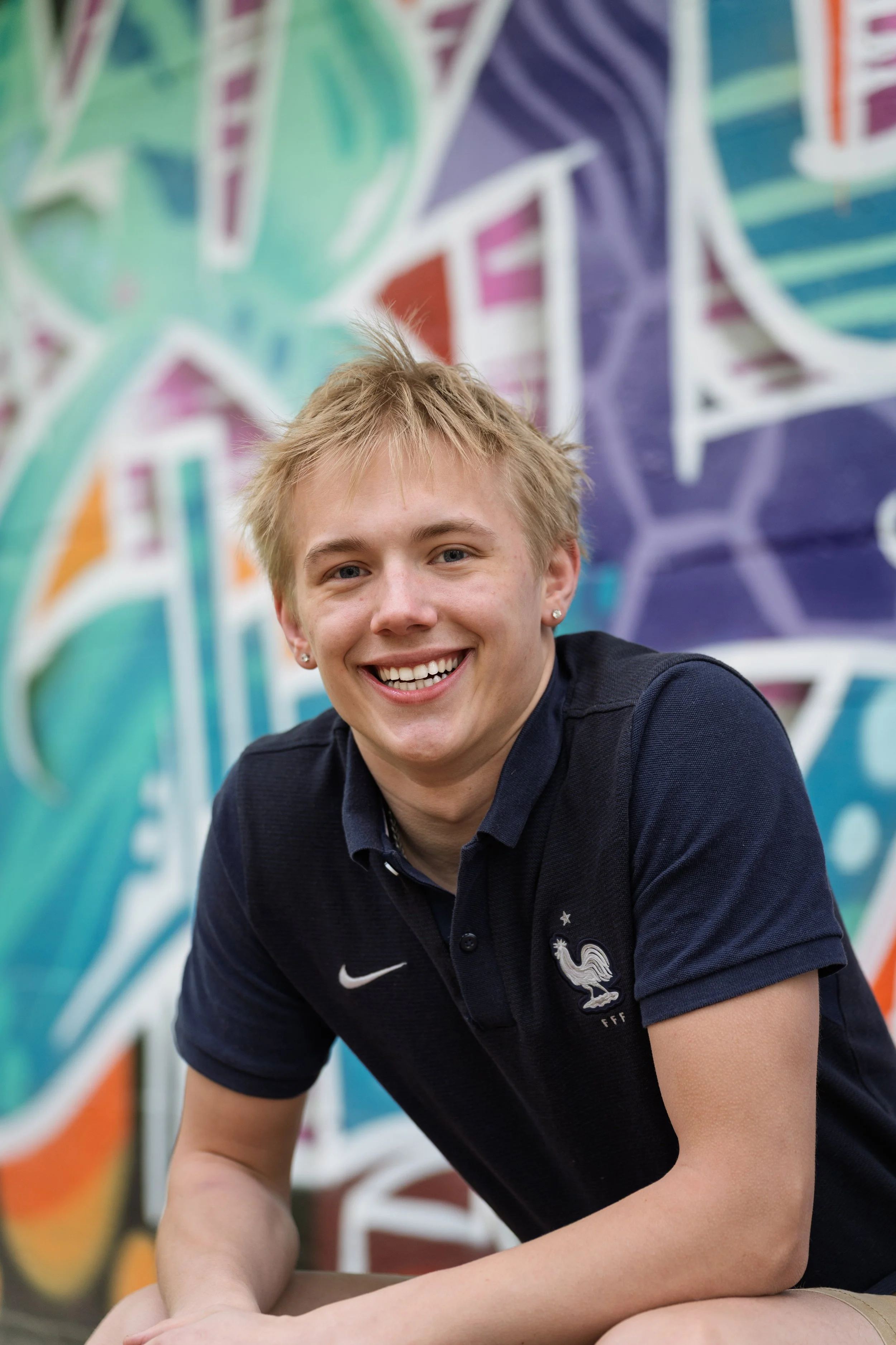 A young man with blond hair smiling, wearing a navy blue Nike polo shirt with a white emblem, sitting in front of colorful graffiti art.