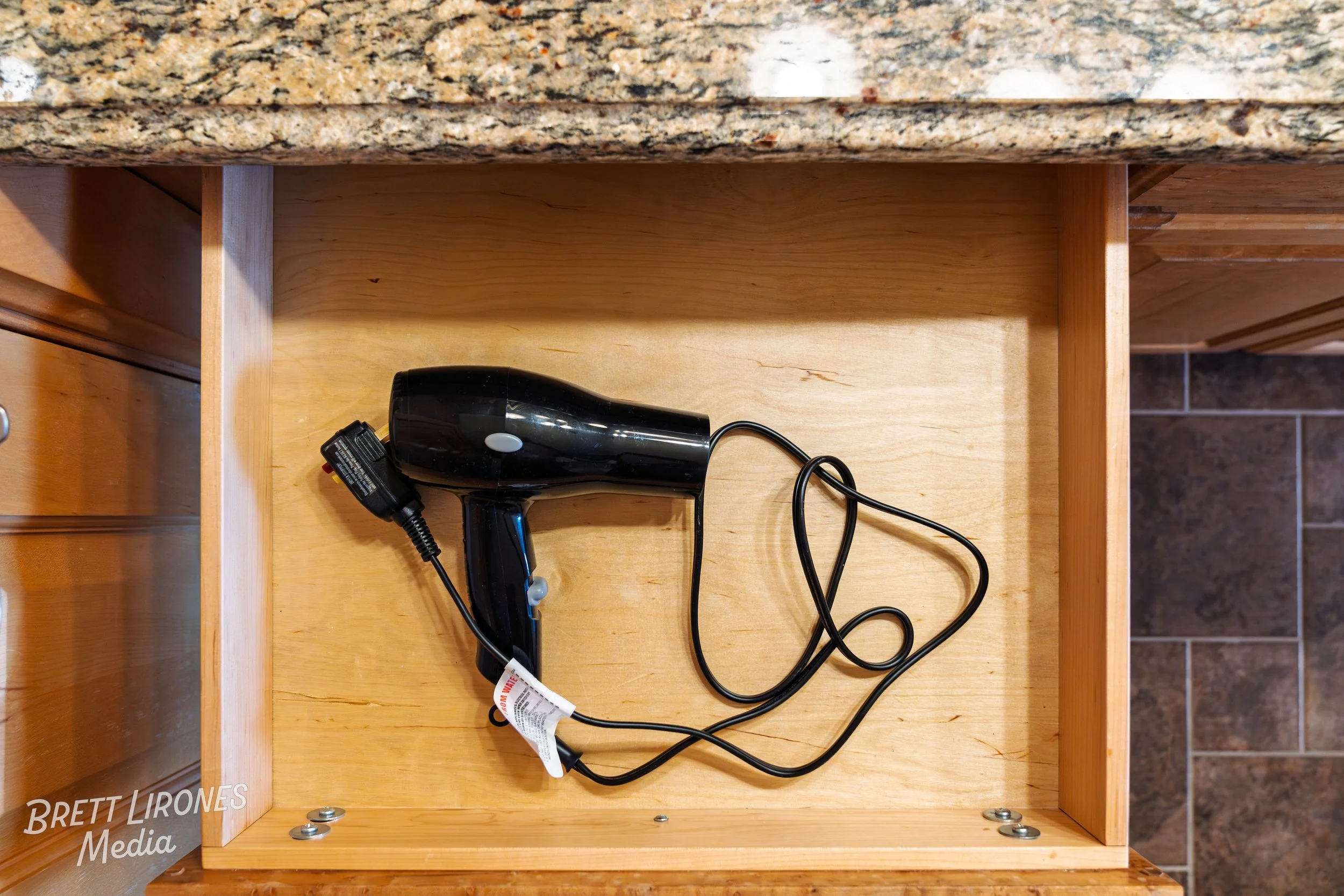 Black handheld hair dryer with cord loosely coiled, inside open kitchen drawer with wooden interior.