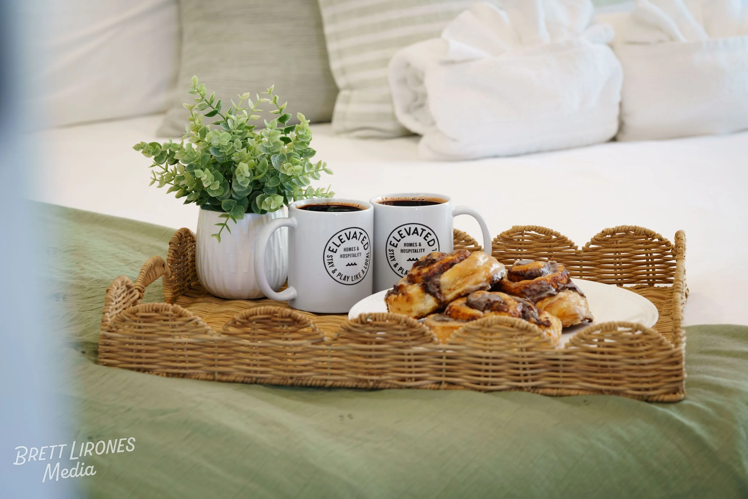 A wicker tray holding two mugs of coffee, a plate of assorted pastries, a potted green plant, and a rolled-up white towel, placed on a table with a bed and pillows in the background.