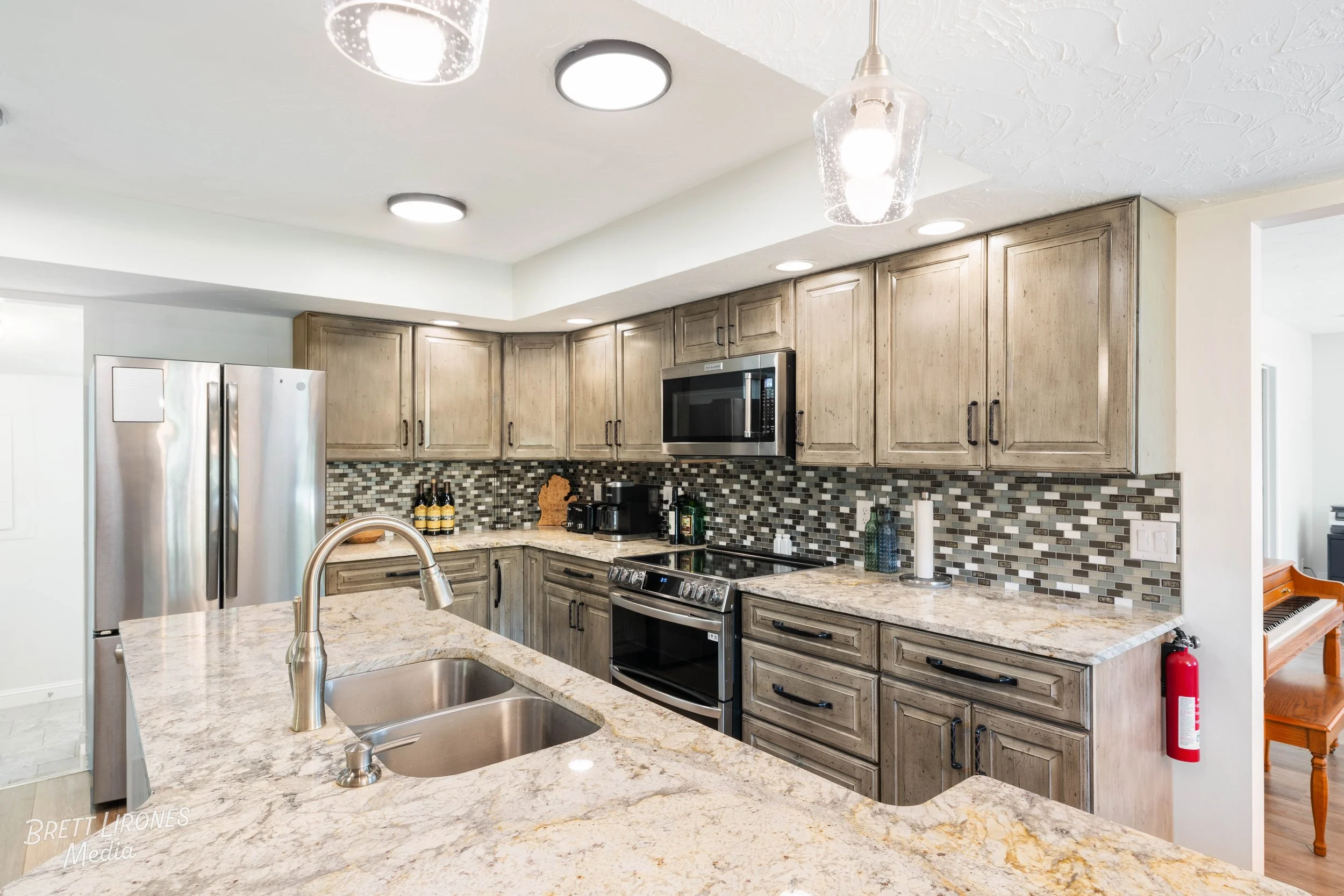 Modern kitchen with granite countertops, light wood cabinets, stainless steel refrigerator, microwave, oven, and mosaic tile backsplash. There are various kitchen items and a fire extinguisher on the wall.