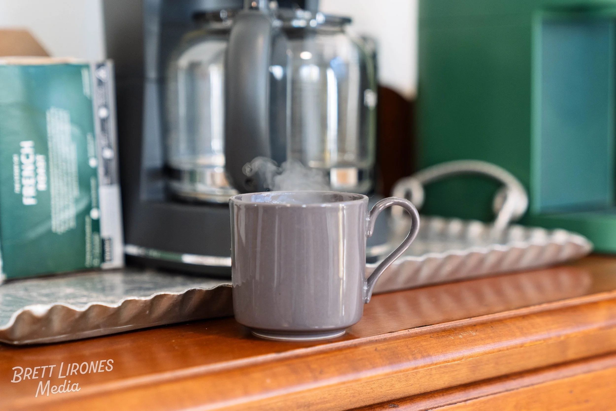 A steaming gray coffee mug on a wooden counter with a coffee machine and a box of tea bags in the background.