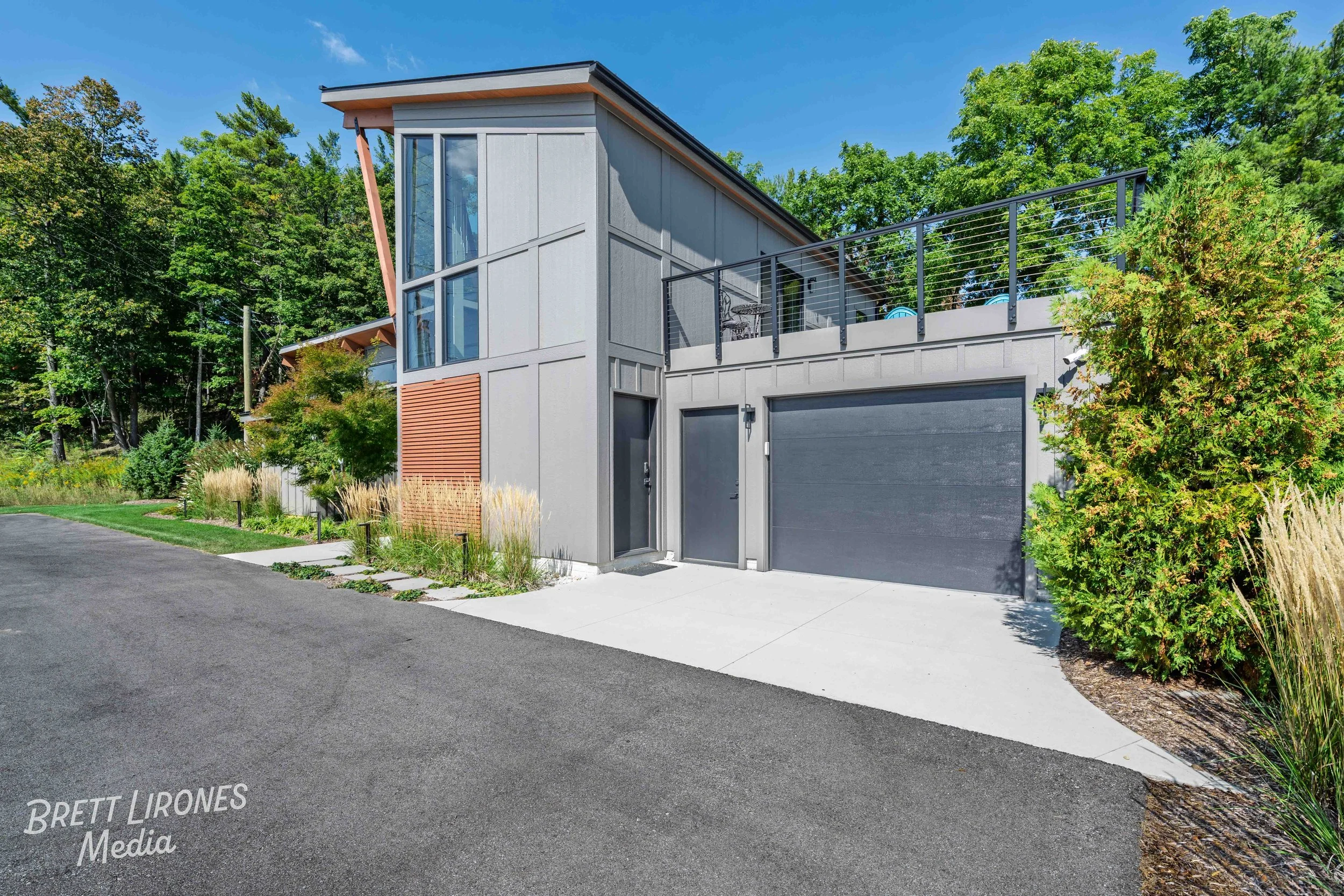 Modern residential house with gray exterior, large windows, a rooftop terrace with furniture, and a garage, surrounded by landscaped greenery and trees.