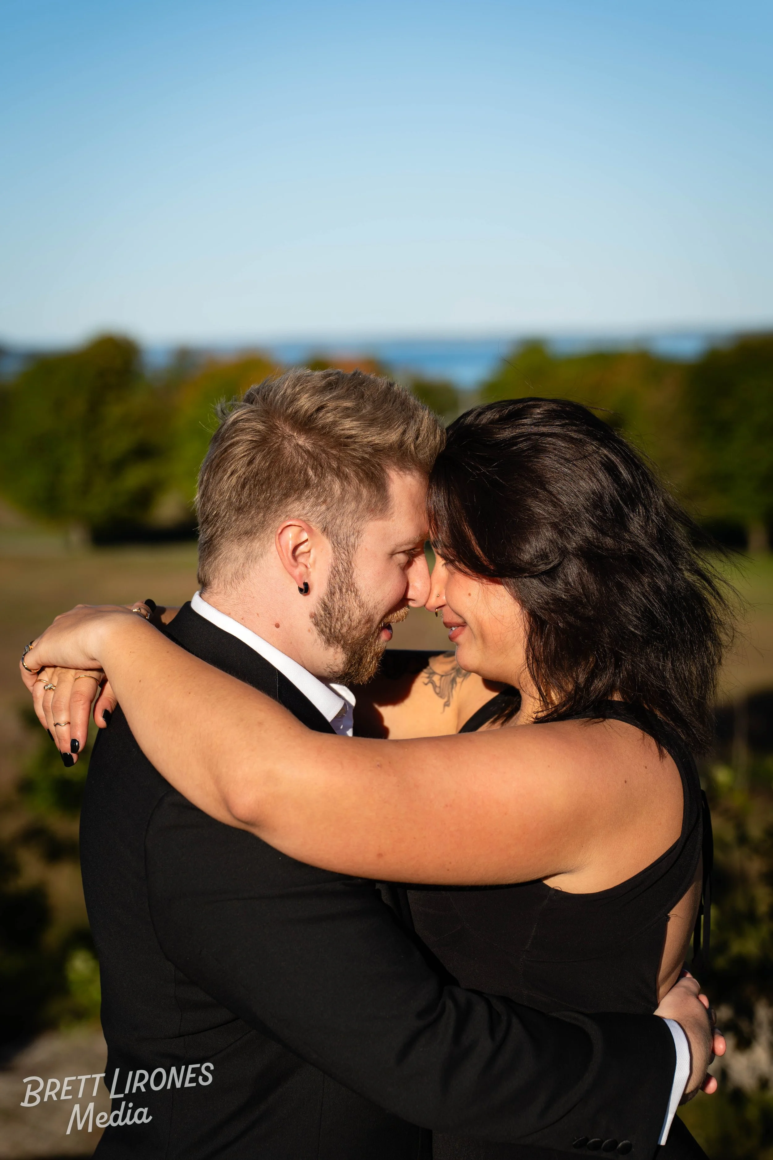 A couple embraces closely outdoors, with their foreheads touching, in a scenic park setting with trees and a blue sky in the background.