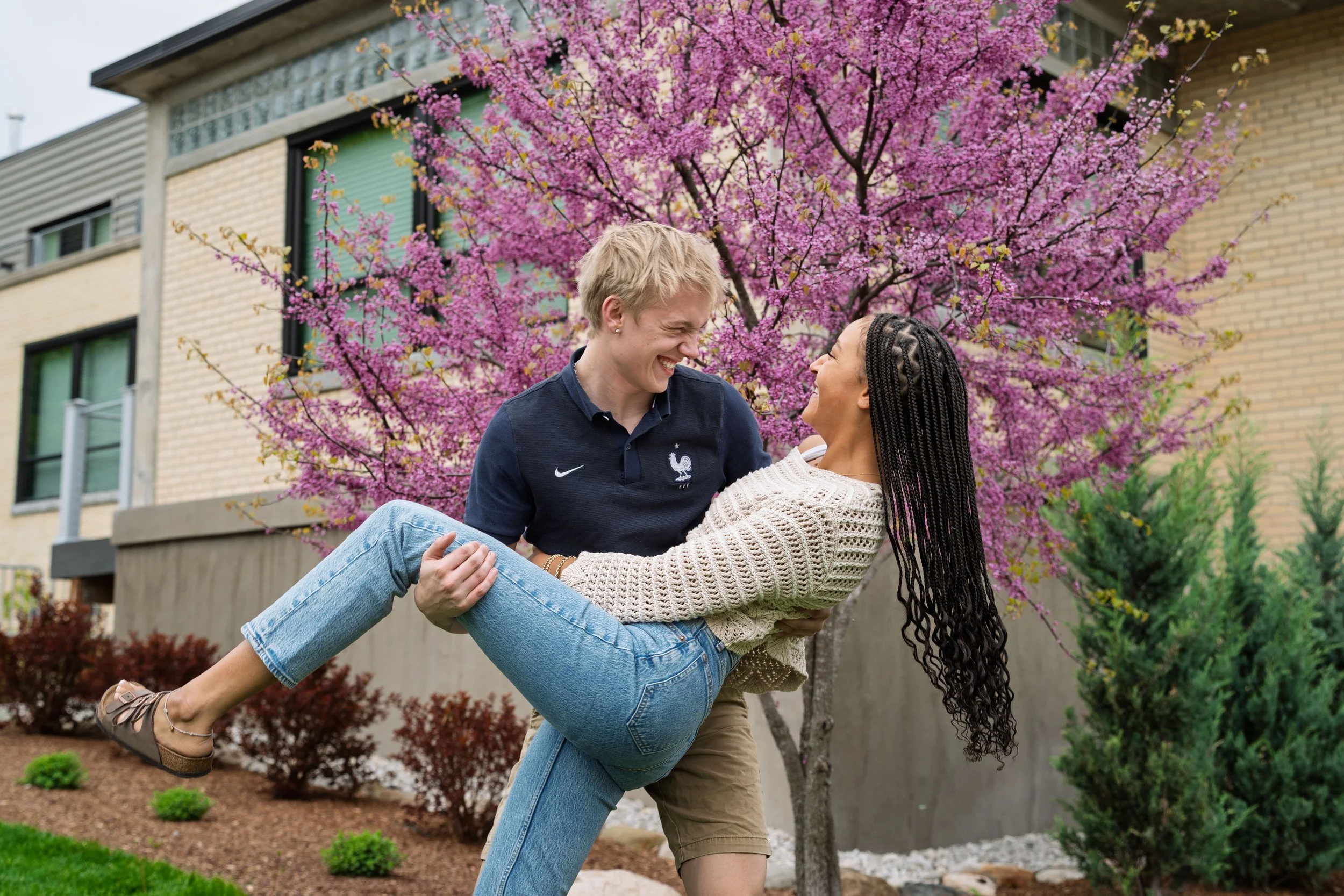 A couple, a man and a woman, are outdoors in a garden, with the man lifting the woman in his arms. They are smiling and looking at each other affectionately, standing in front of a pink flowering tree and a building with brick and glass windows.